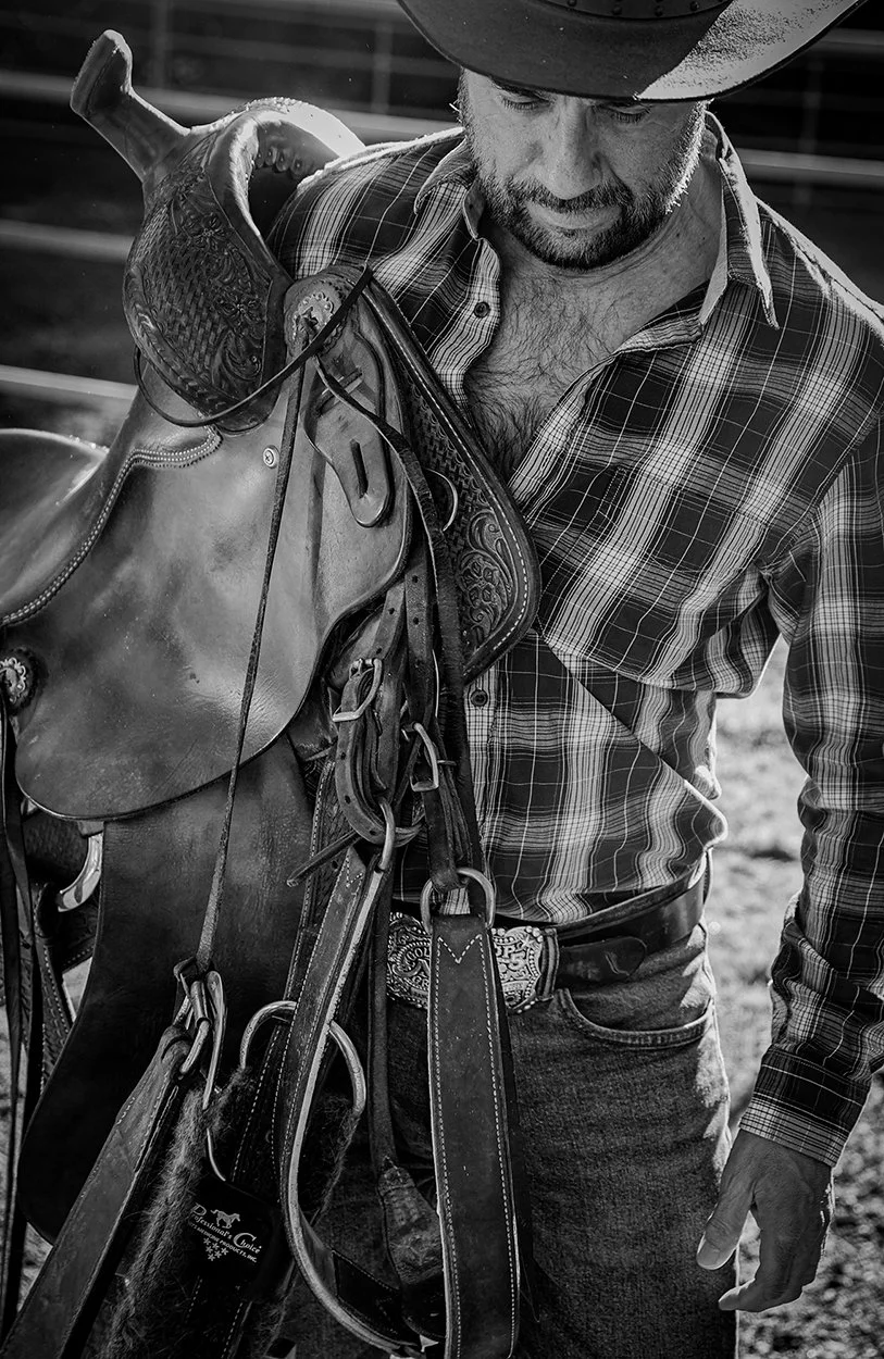 This is a striking black and white photograph capturing a rugged cowboy in a moment of preparation. The man, with a beard and a dark cowboy hat, is carrying a Western saddle and various pieces of leather tack, which appear to be bridles and reins, sl