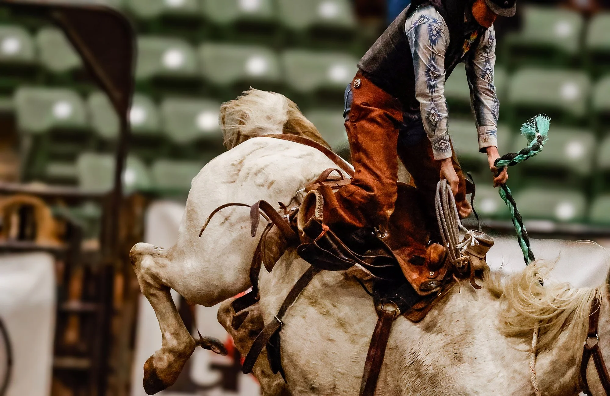 A high-intensity saddle bronc riding moment showing a bucking horse mid-kick in an indoor rodeo arena. The rider is gripping the bronc rein, wearing chaps, a long-sleeve patterned shirt, and rodeo gear. Arena seating is blurred in the background, emp