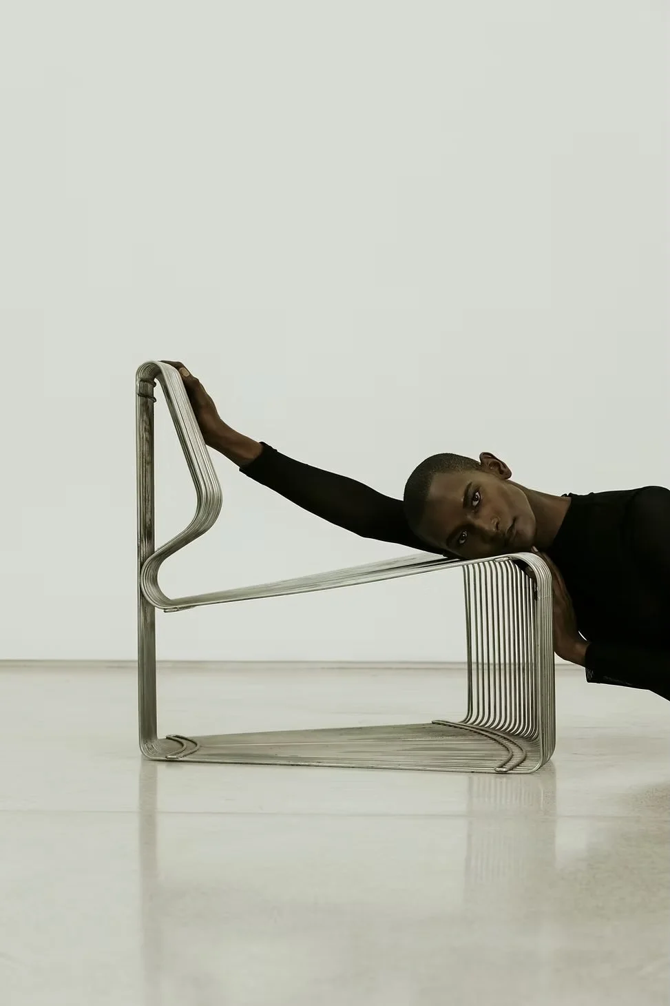 A person leaning on a modern metal chair with an abstract, continuous design, resting their head and arm on it, in a minimalist setting.