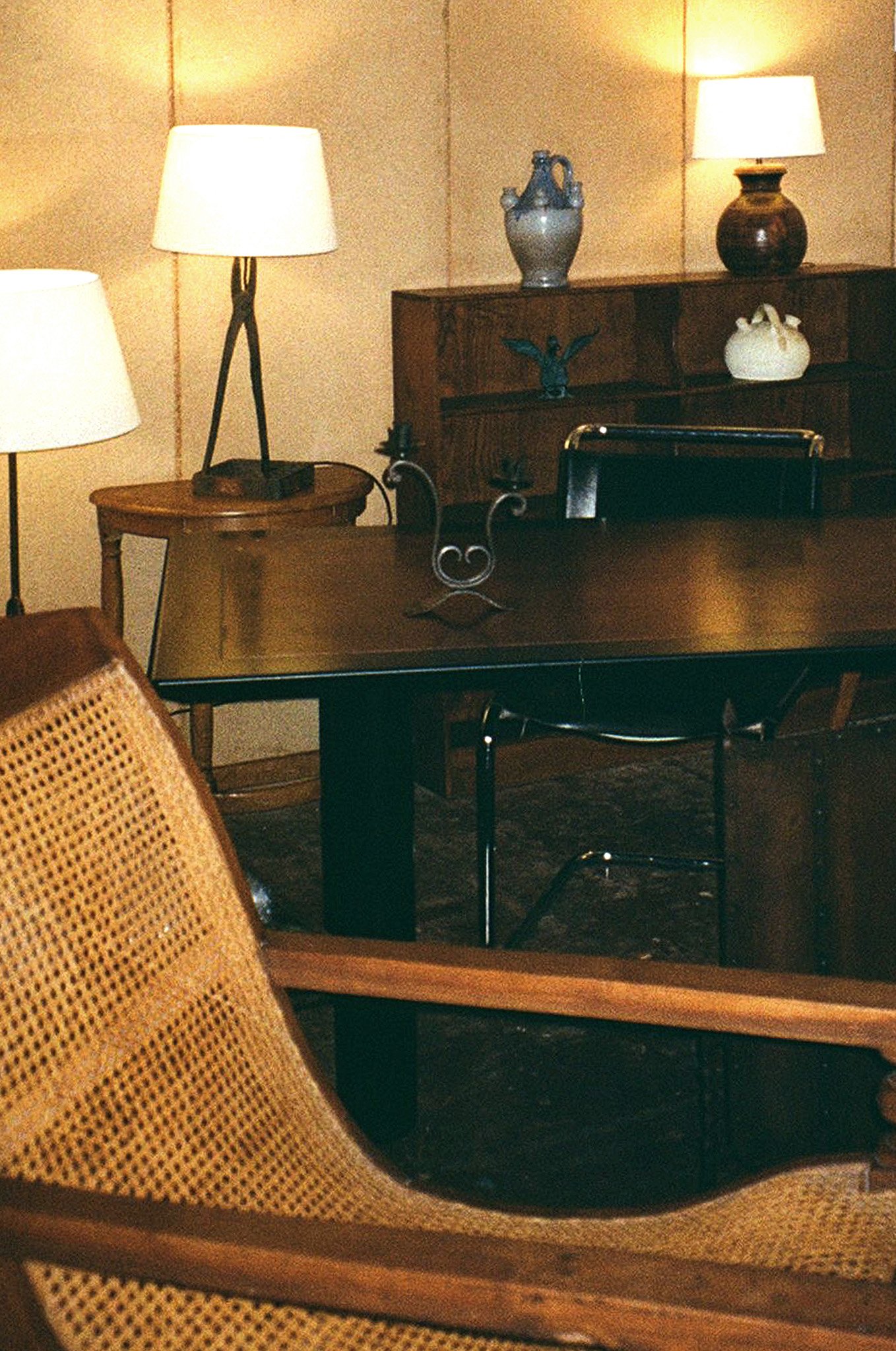 Living room with wooden furniture, table, and shelves. The shelves hold decorative vases and figurines. There are traditional lamps on the shelf and a round side table. Part of a netted or woven furniture piece is visible in the foreground.