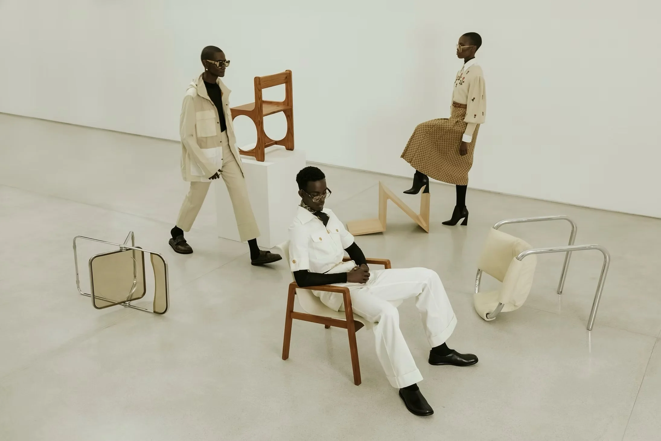 Four people with dark skin and glasses in a minimal white art gallery. One person in a white jumpsuit with black boots sits on a wooden chair. Two people, one standing with beige pants and a vest, another sitting with a plaid skirt and high heels, are guiding the scene. The fourth person standing near a wooden chair is looking forward. Art pieces include a wooden sculpture and various chairs on the floor and against the wall.