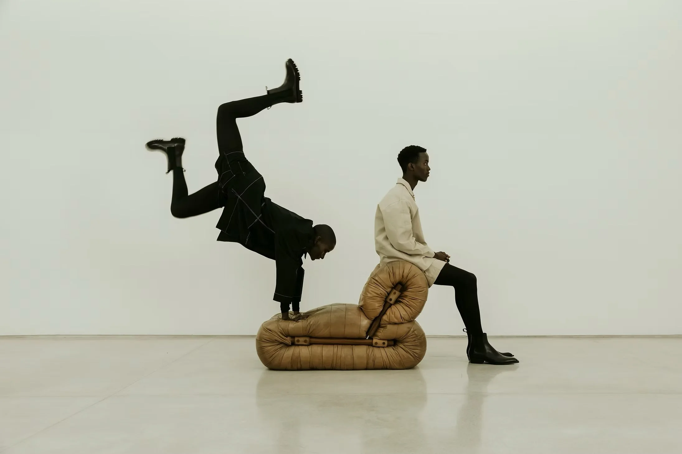 Two people sit on a padded, cushion-like seat, with one person sitting calmly and the other person doing a handstand on the back of the seat.