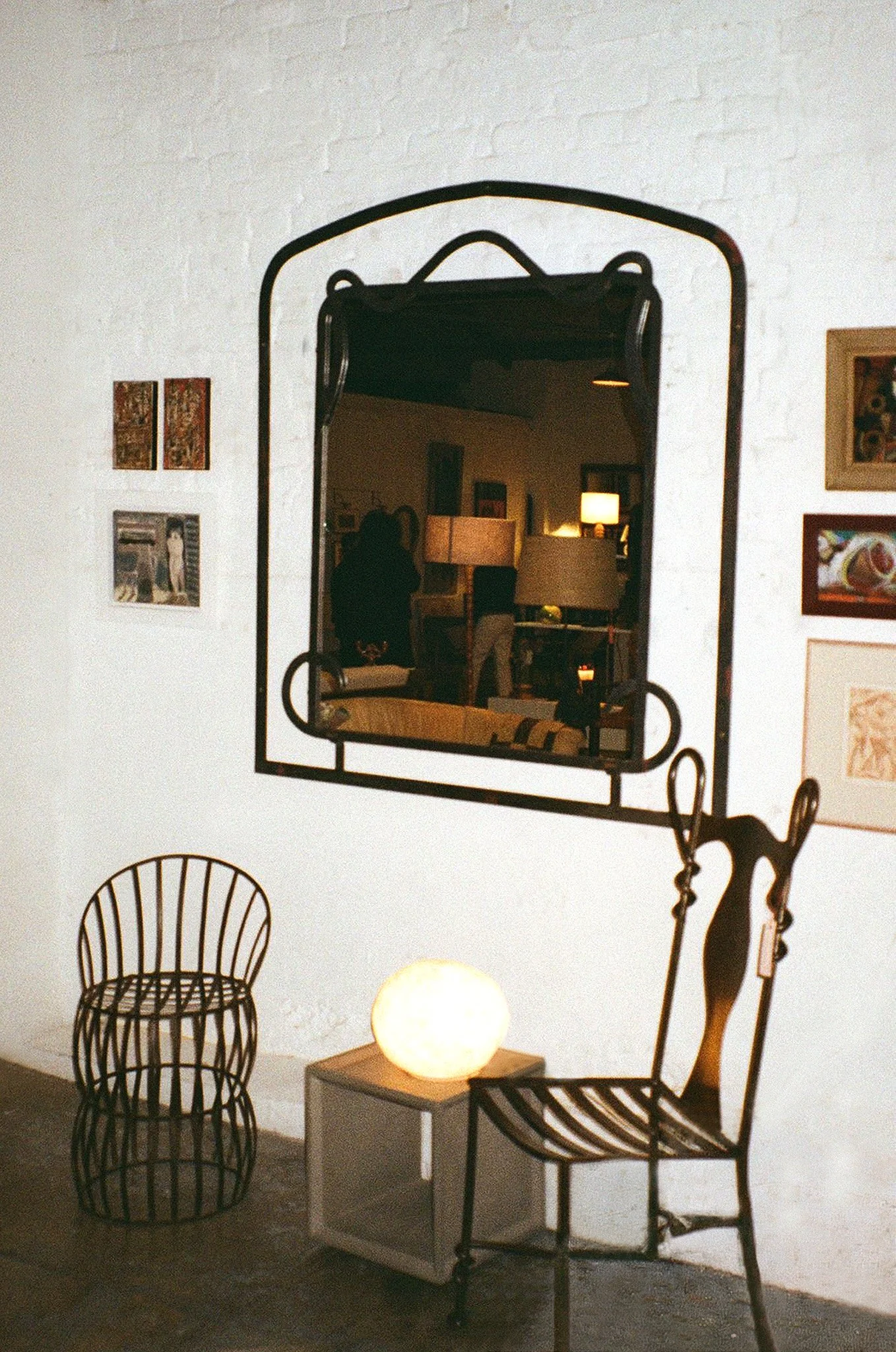 Interior of an art gallery or museum with wall-mounted artwork, wireframe chairs, a glowing spherical lamp on a small table, and a mirror reflecting a person taking a photo.