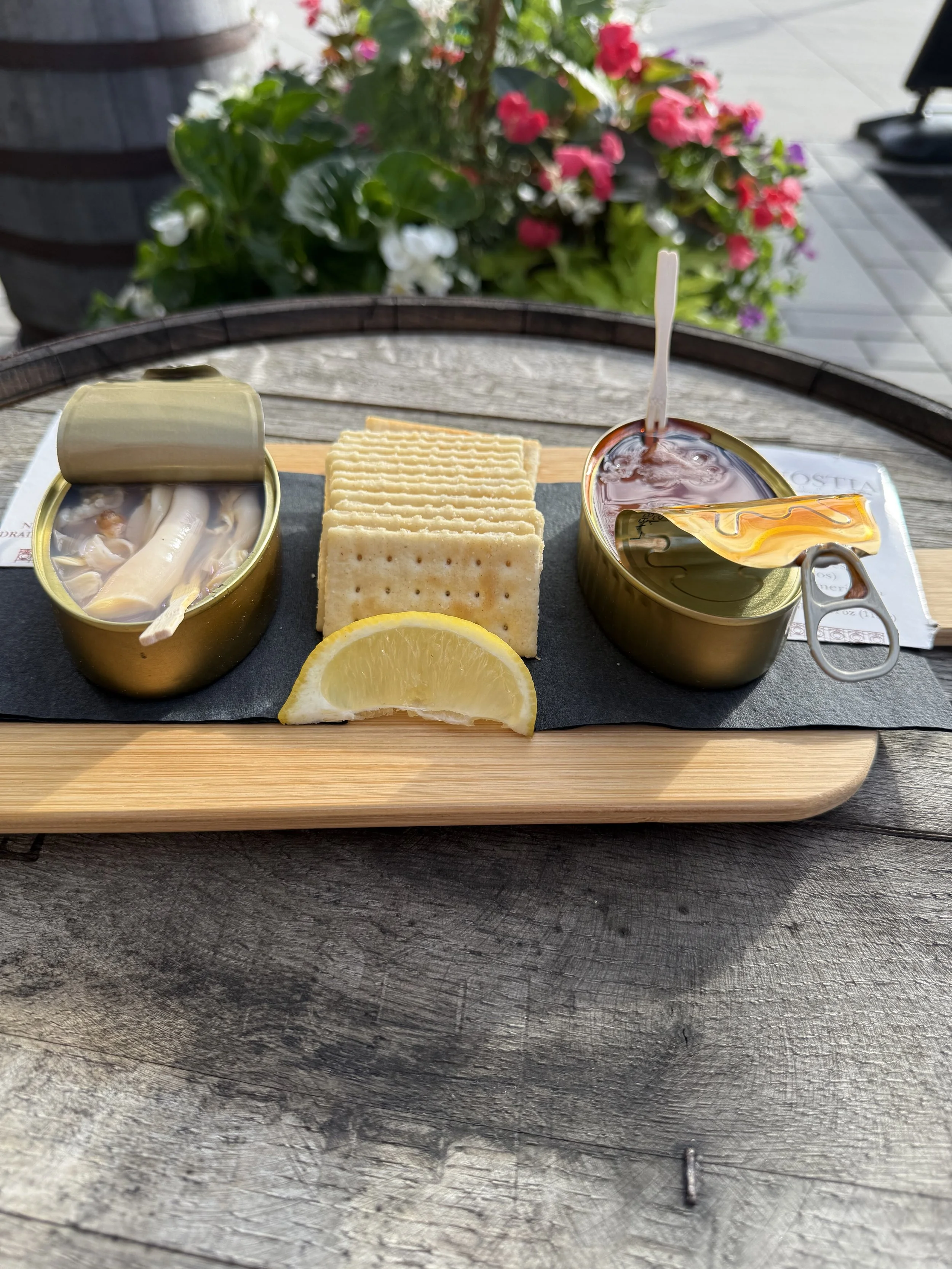 Can of canned clams, Ritz crackers, lemon wedge, and a bowl of clam chowder on a wooden tray outside with greenery and flowers in the background.