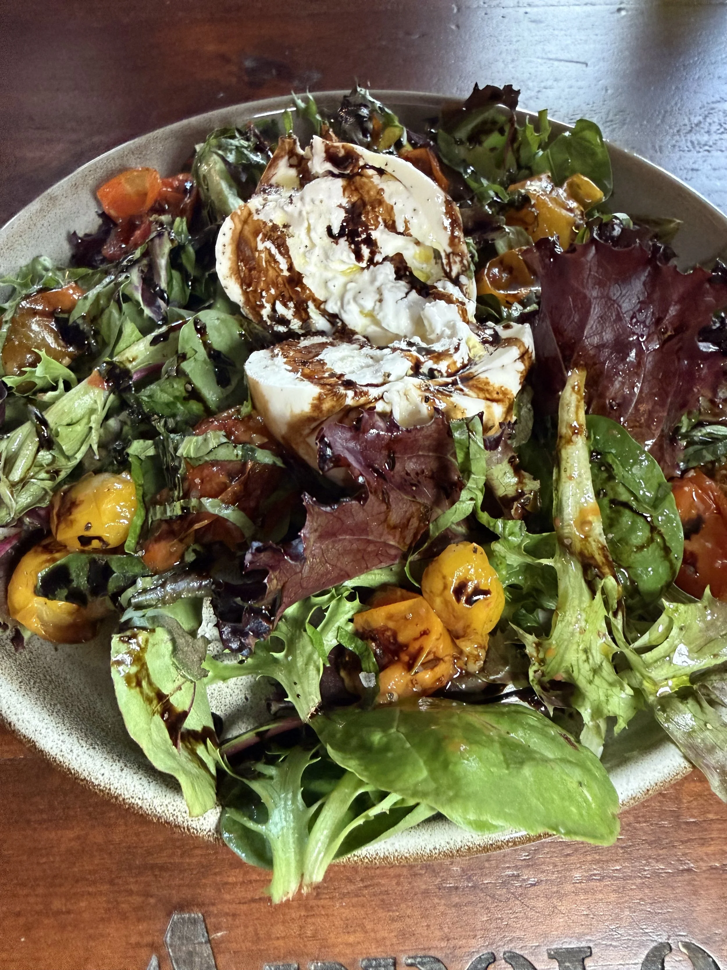 Mixed green salad with cherry tomatoes, leafy greens, and a dollop of burrata cheese drizzled with balsamic reduction on a ceramic plate.