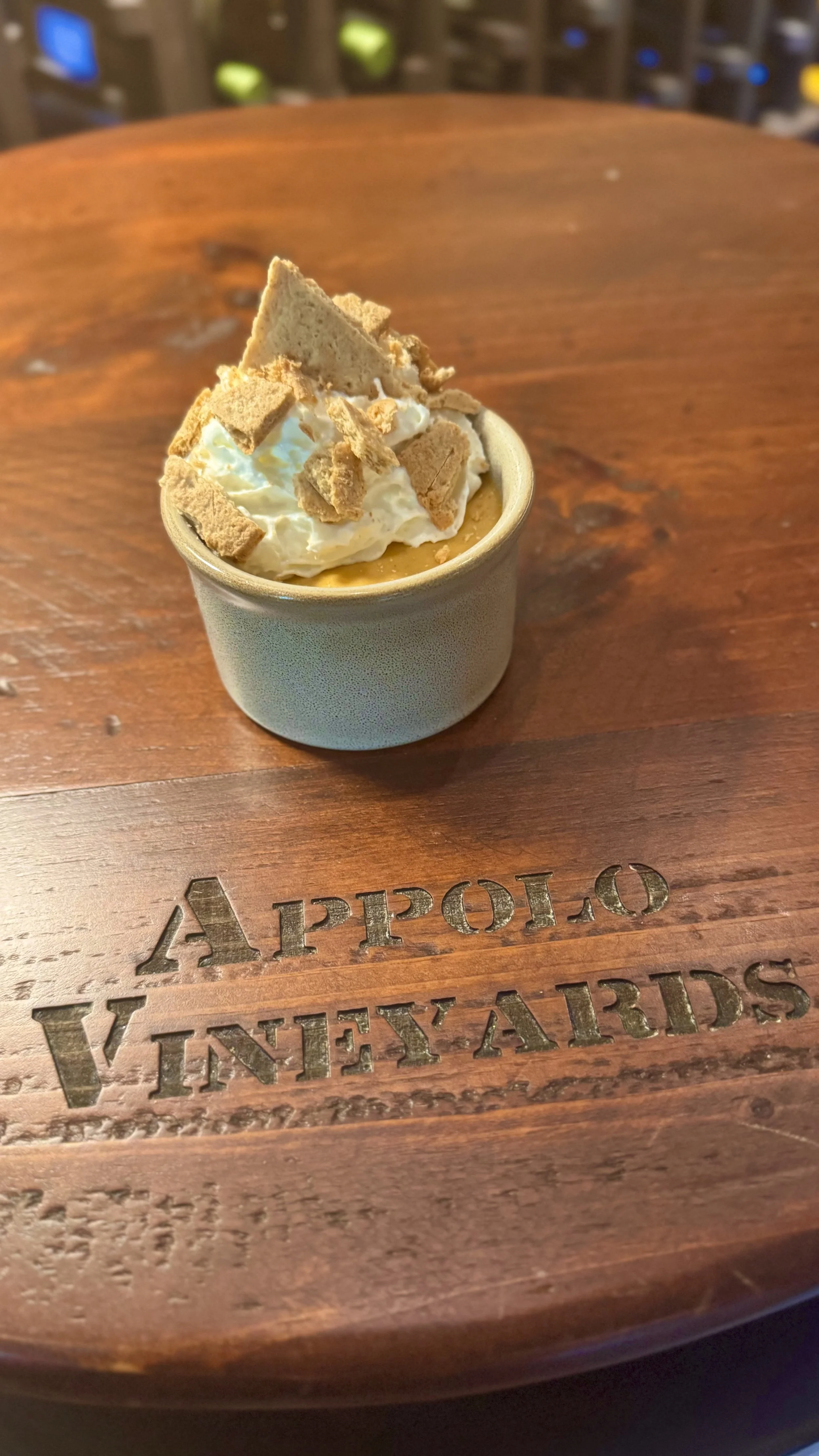 A cup with whipped cream and crushed cookies on top, placed on a wooden tray with engraved text.