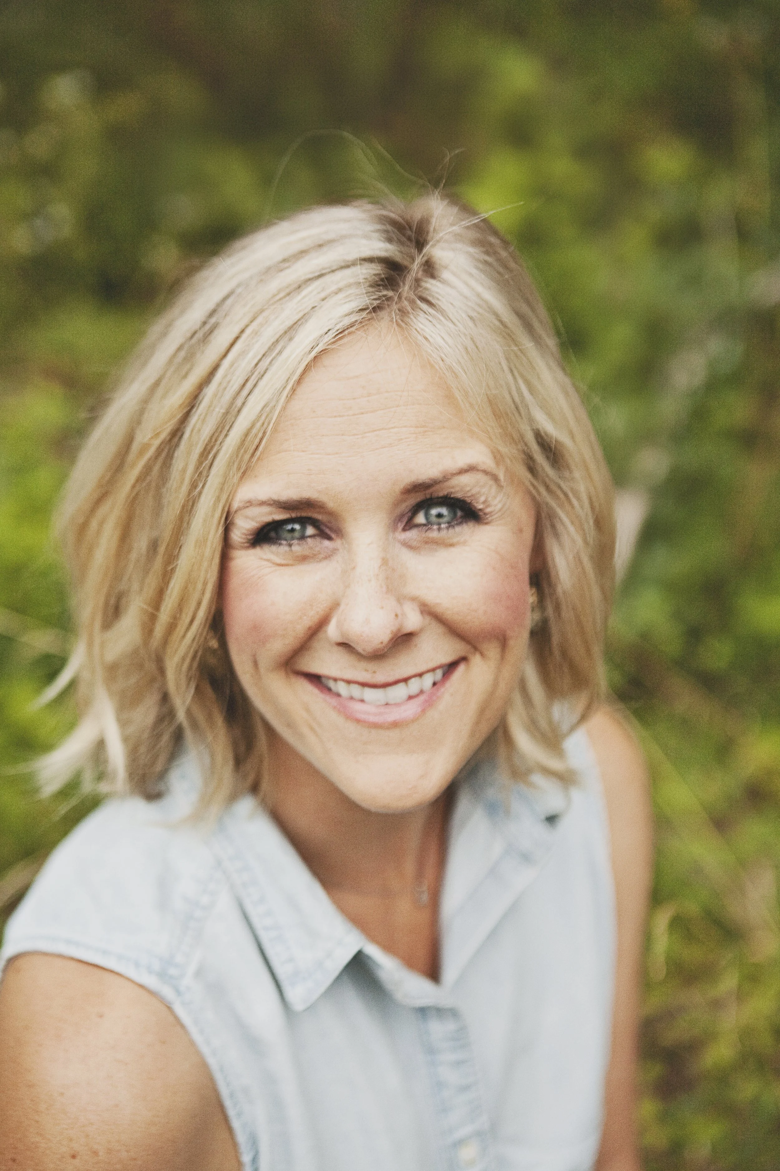 A woman with blonde hair smiling outdoors, wearing a sleeveless denim shirt, with a blurred green background.