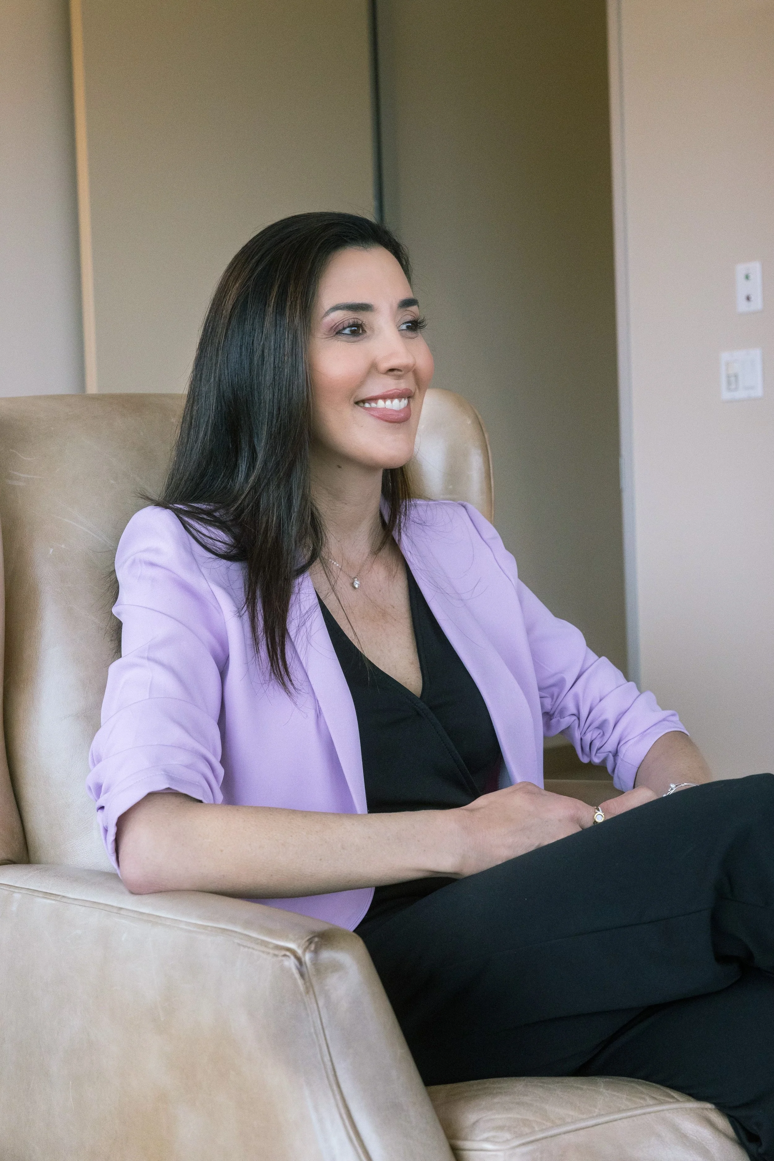 A woman with long dark hair, wearing a black top and a light purple blazer, sitting in a beige chair and smiling.