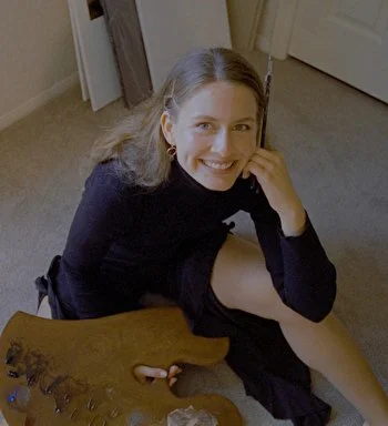 A woman with long blonde hair sitting on the floor, smiling, wearing a black top and black skirt, in a room with beige carpet and white walls.