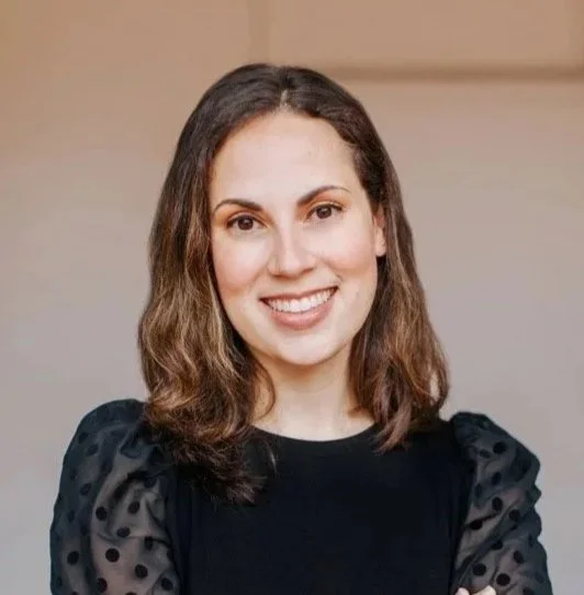 A woman with shoulder-length curly brown hair, smiling, wearing a black top with sheer polka-dot sleeves, standing indoors with a neutral background.