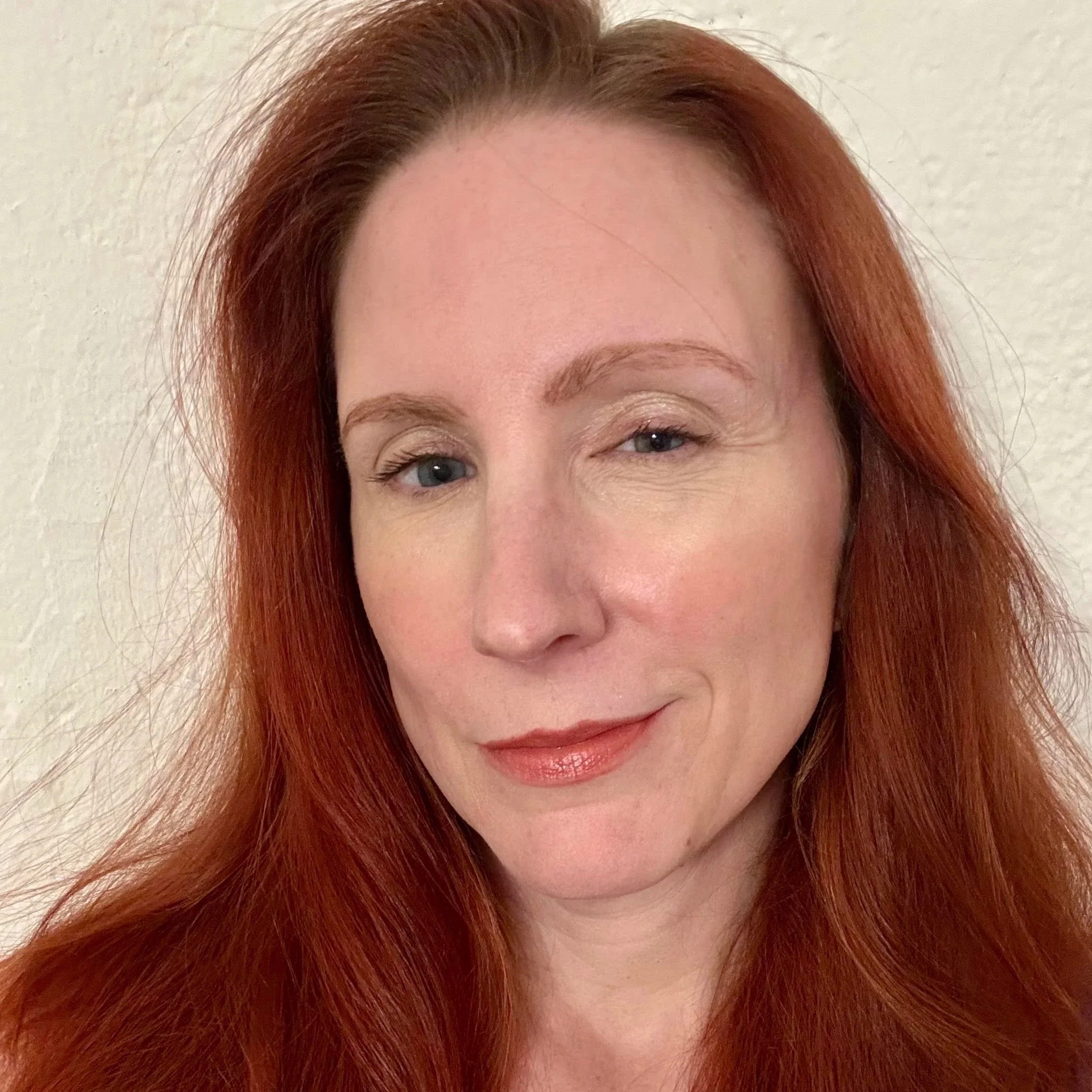 Close-up of a smiling woman with long red hair, wearing makeup and a dark top, against a neutral background.