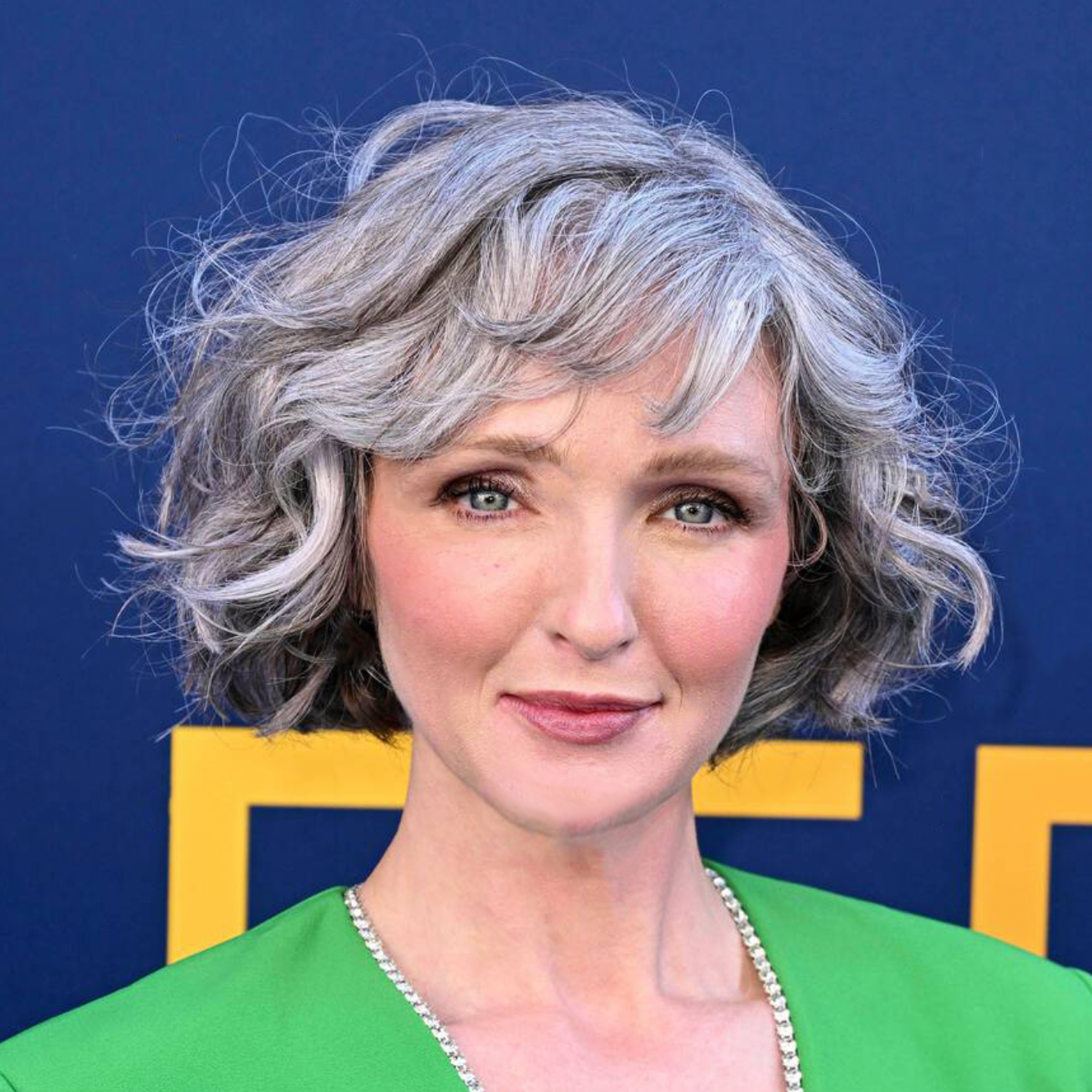 Portrait of a middle-aged woman with short, curly gray hair and blue eyes, wearing a bright green top and a delicate necklace, with a neutral background.