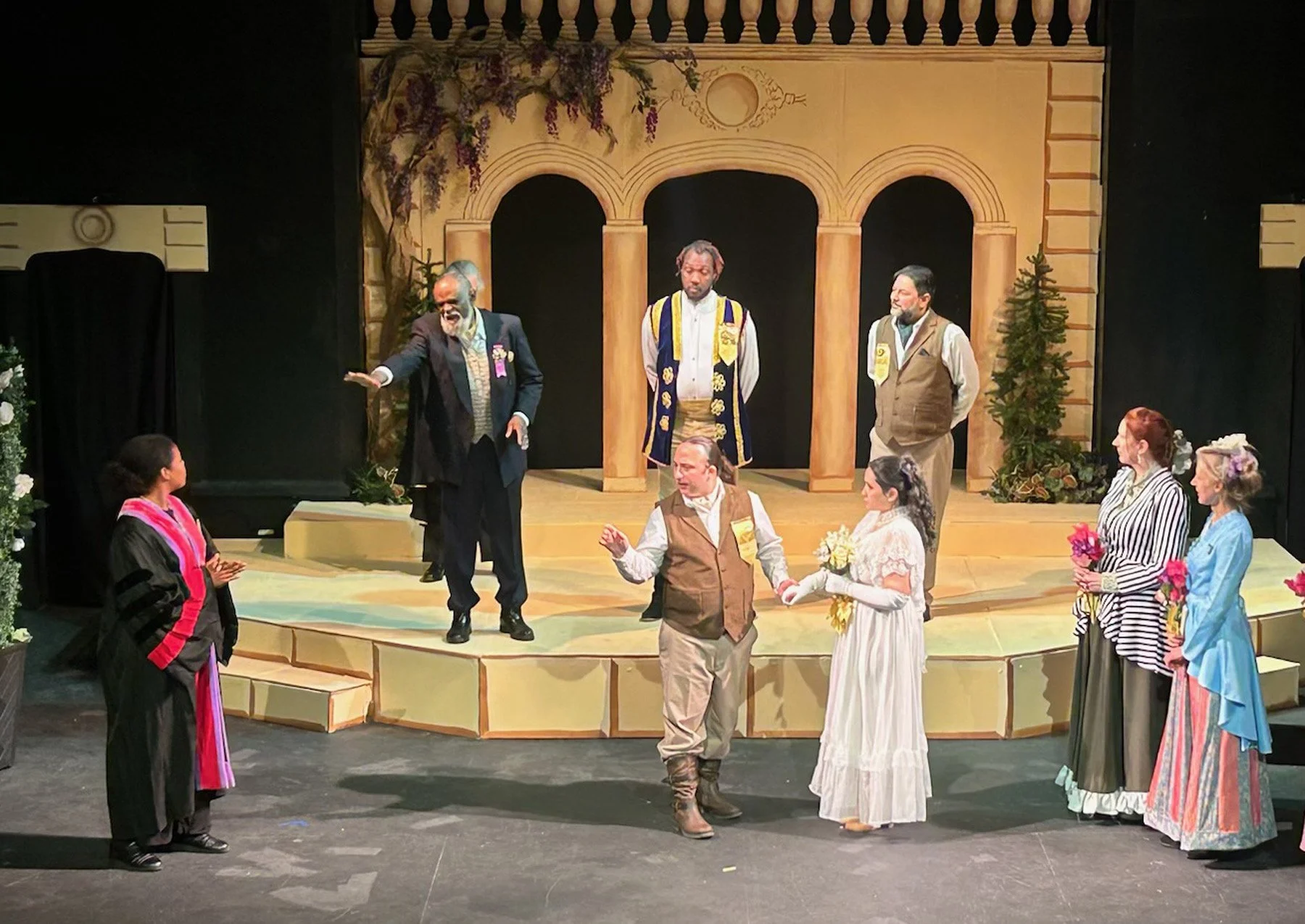 Actors on stage performing a play set in a garden with a stone archway background, dressed in period costumes, some holding flowers, engaged in dialogue.