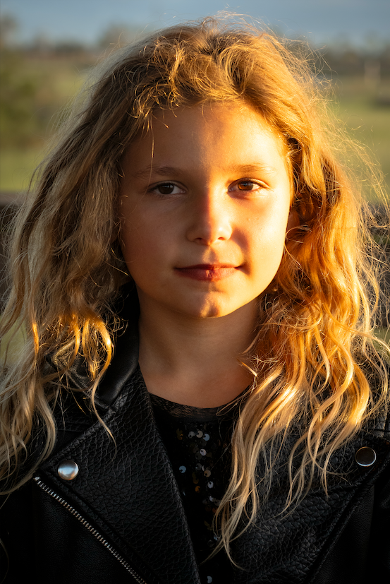 A young girl with long, curly blond hair standing outdoors in sunlight, wearing a black leather jacket.