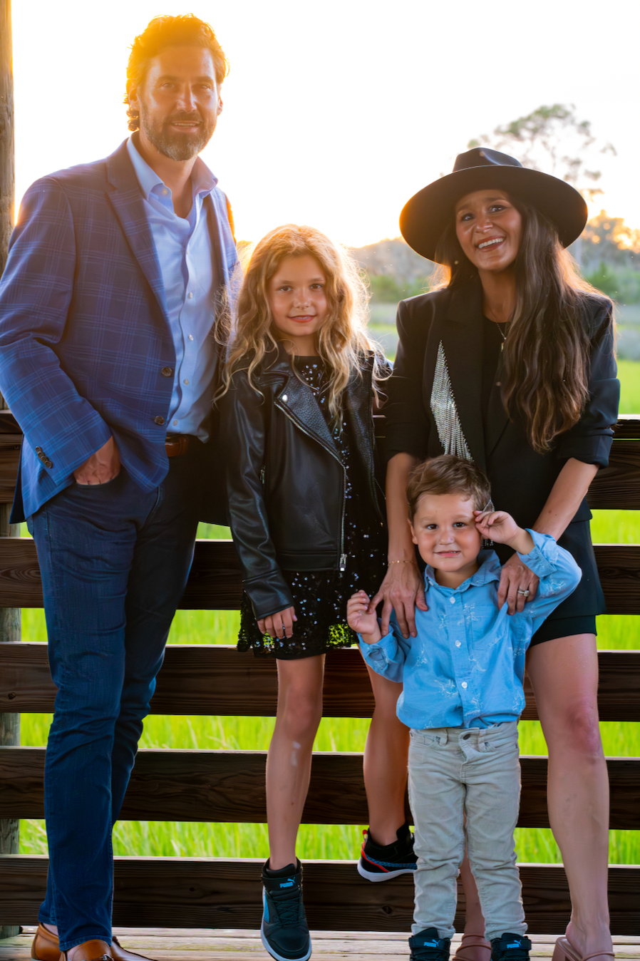 A family of five posing outdoors on a wooden bridge during sunset, with green fields in the background. The father is wearing a blue checkered blazer and jeans, the mother has long dark hair and wears a black hat and blazer, the daughter has curly blonde hair and is dressed in a leather jacket and dress, and the young son is wearing a light blue shirt.