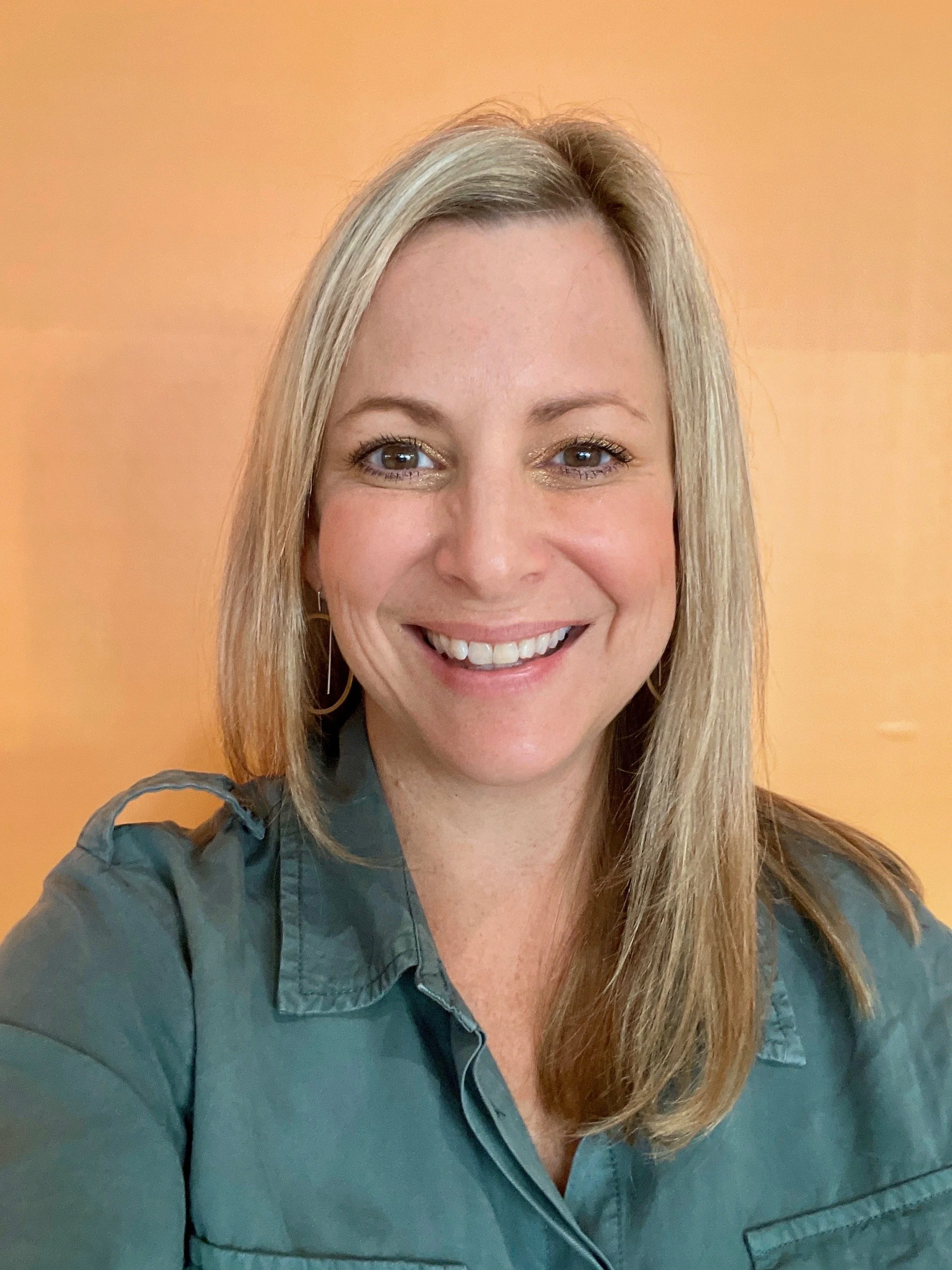 A smiling woman with shoulder-length blonde hair wearing leopard print glasses and a white blouse with small black bird prints, taking a selfie indoors.