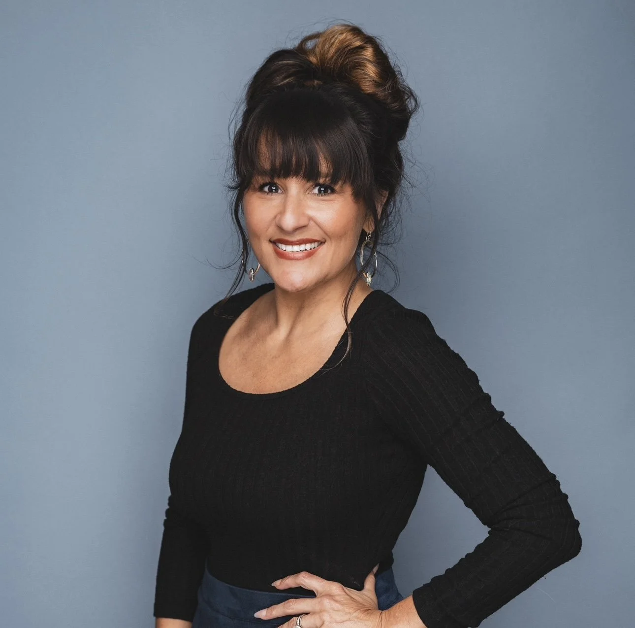 A woman with dark hair styled in a high bun with blonde highlights, wearing a black long-sleeve top and earrings, standing against a blue background, smiling at the camera.