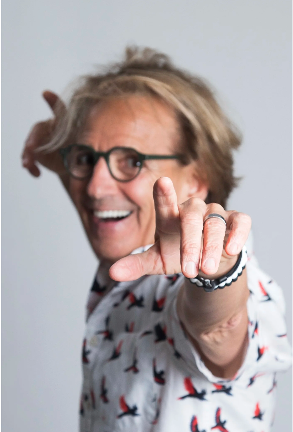A man with glasses and light brown hair smiling and pointing at the camera, wearing a shirt with a bird pattern.