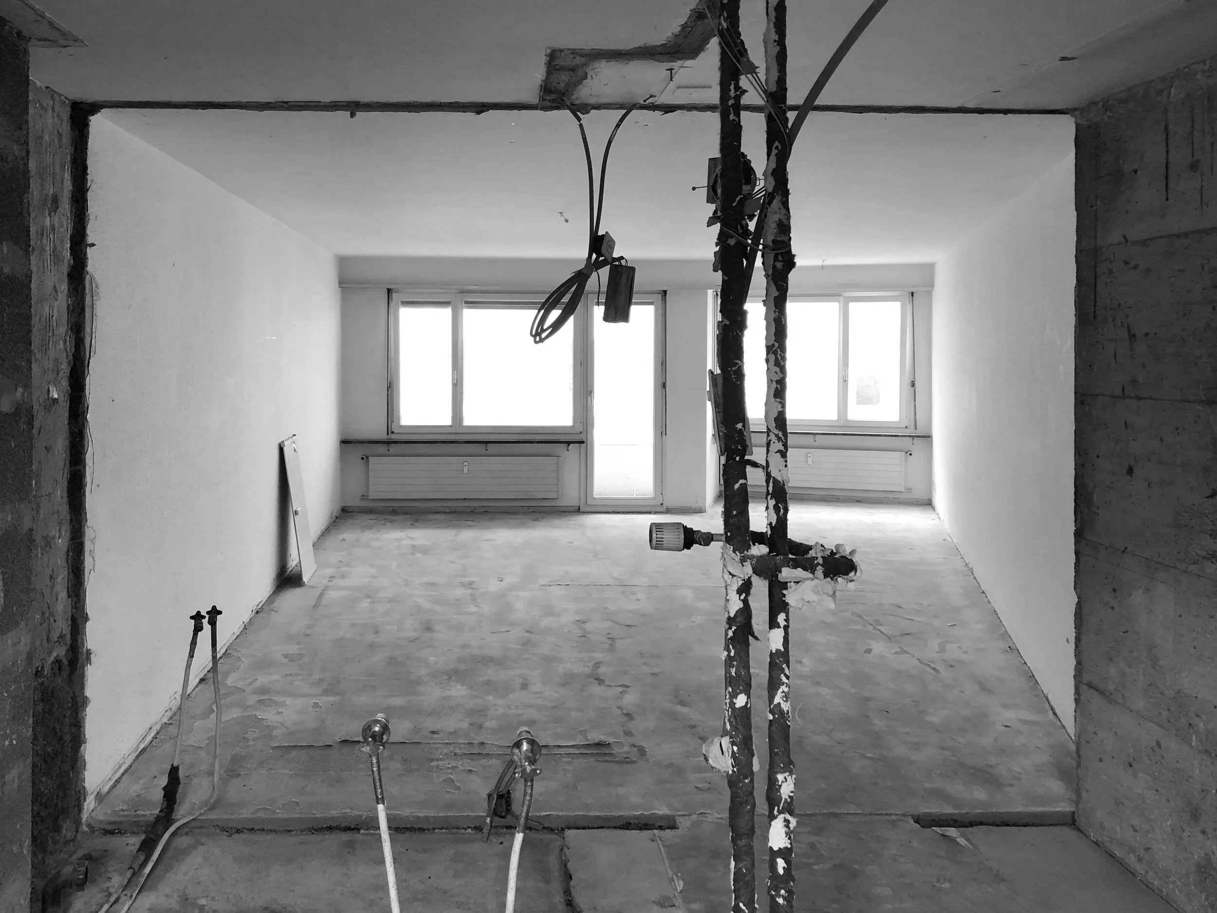 Leerer Raum in einer Wohnung unter Renovierung, mit großen Fenstern und sichtbarer Rohrleitung im Vordergrund.
