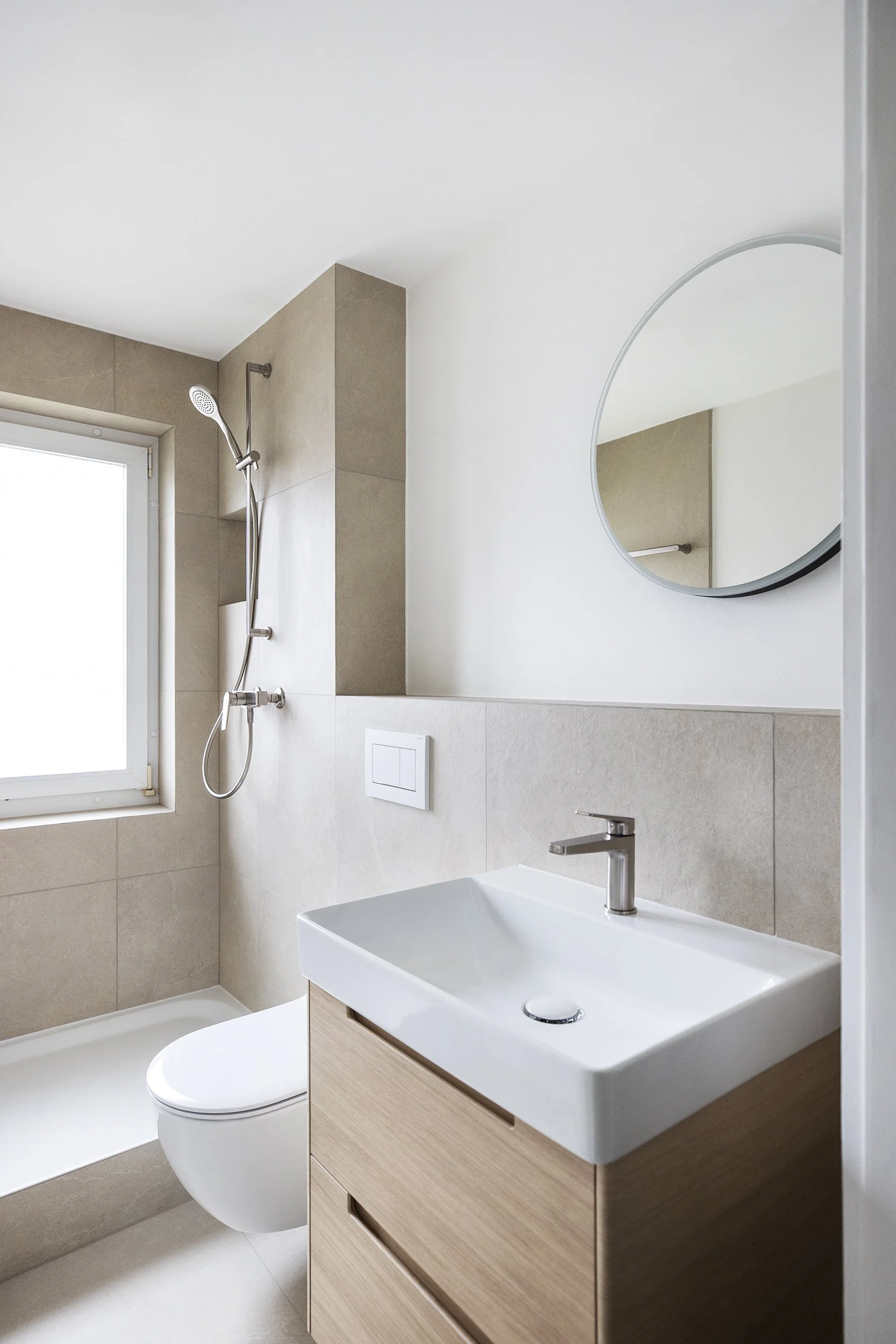 Ein modernes Badezimmer mit einer weißen Wand, einem runden Spiegel, einem Holzwaschbecken und einer Toilette. Im Hintergrund ist eine Dusche mit einem Fenster.