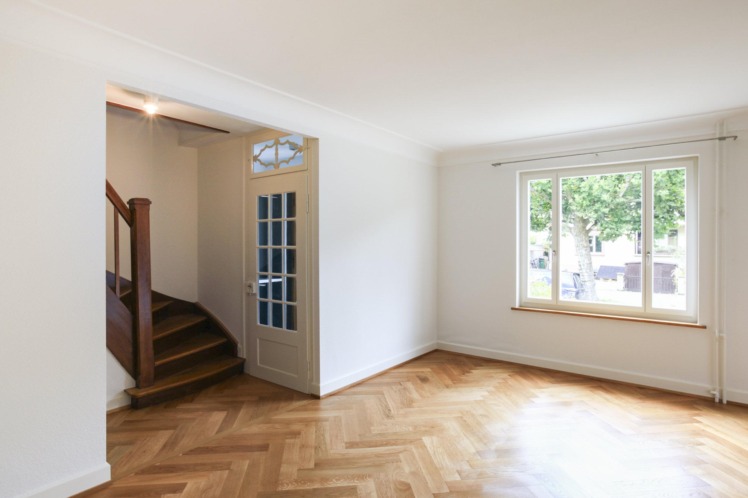 Helles, unmöbliertes Wohnzimmer mit Holzboden, großem Fenster und Treppe am Eingang