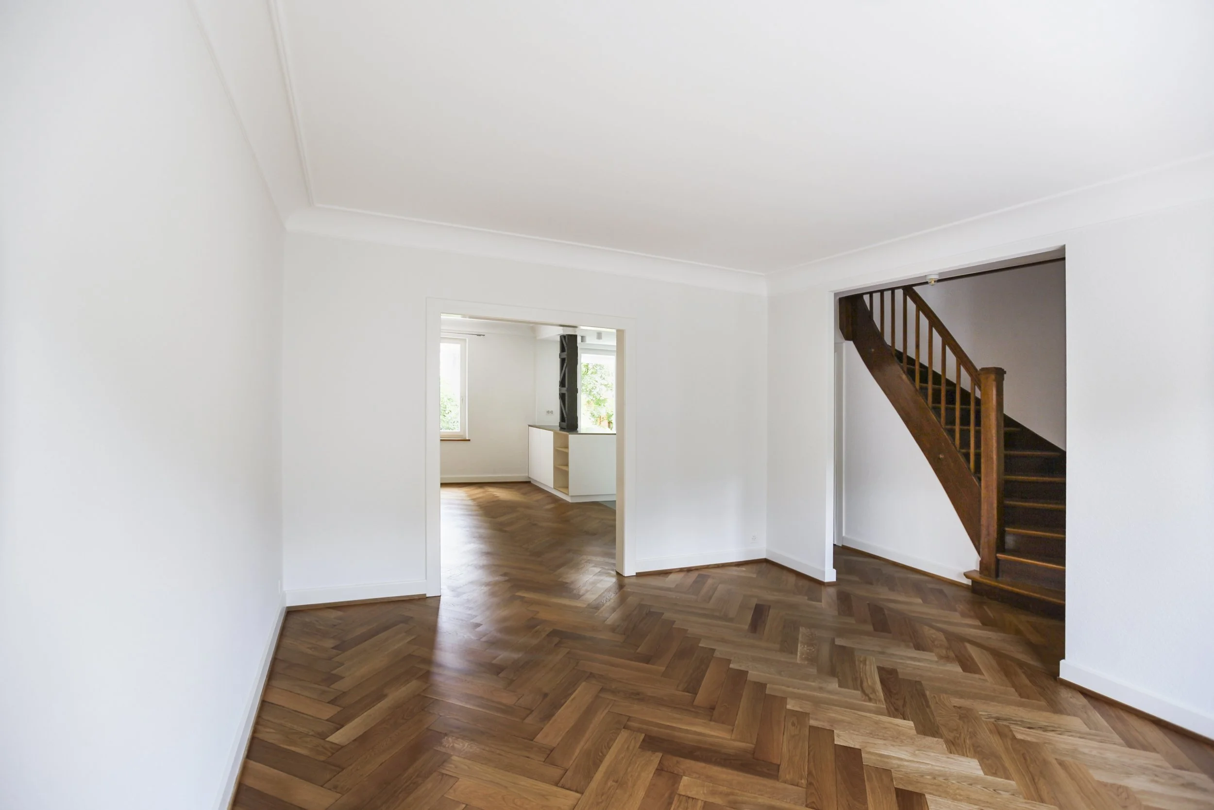 Leeres wohnzimmer mit weißen Wänden, hölzernem Parkettboden in Fischgrätmuster, sichtbare Treppe aus Holz und Durchgang zu einem weiteren Raum mit Fenster.