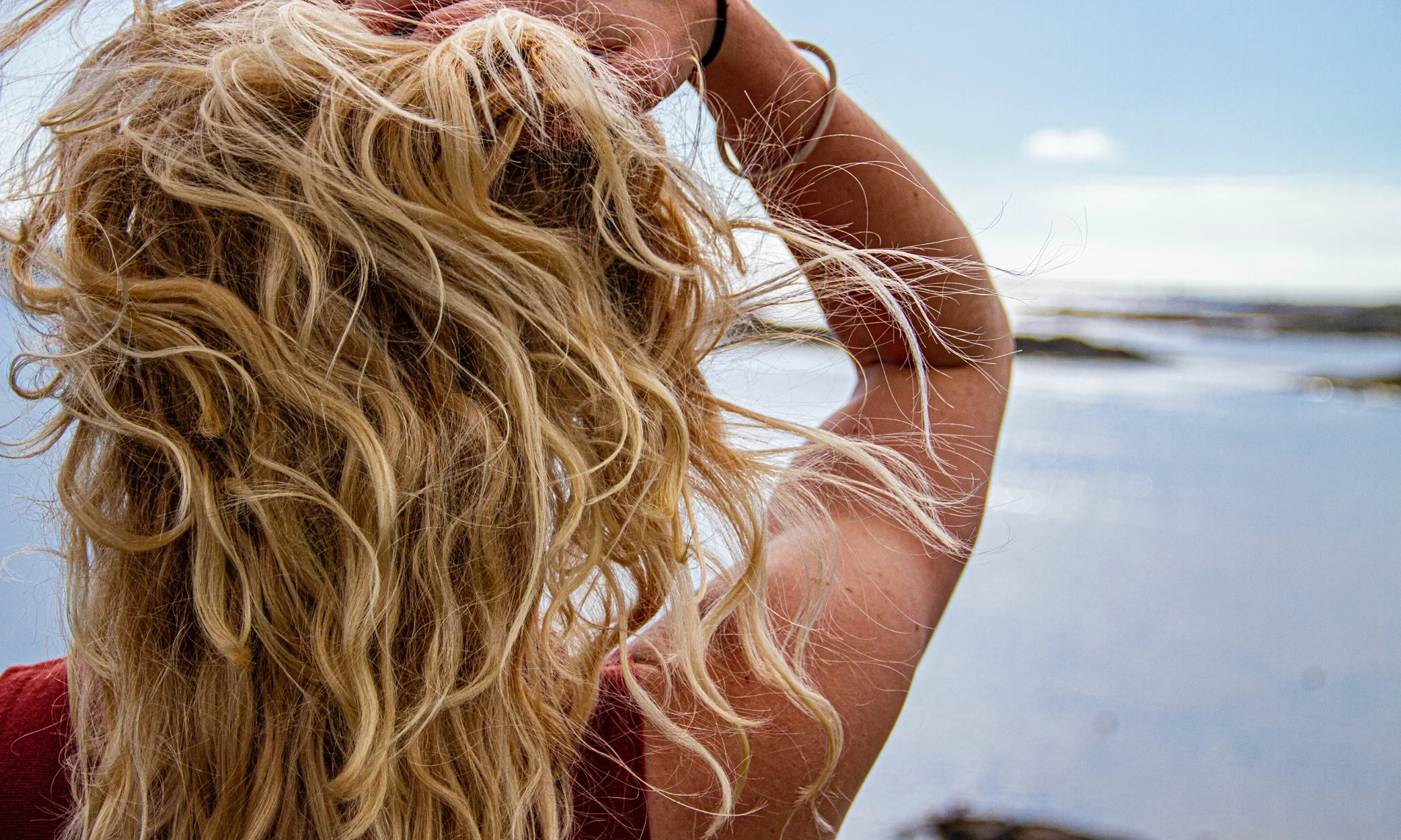 A woman with long, curly blonde hair standing near a body of water, with her hand resting on her head and her face turned away.