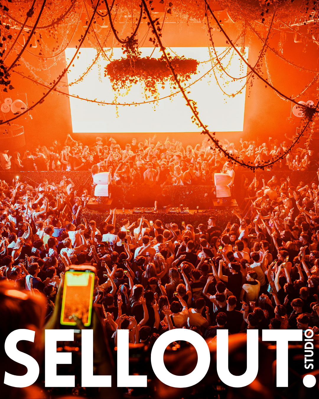 Crowd of people dancing and enjoying a DJ performance at a music event, with stage lighting and hanging decorations.