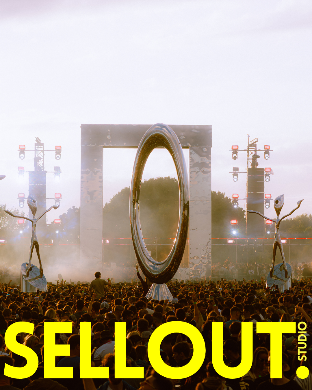 Large crowd at outdoor music festival with stage, large metallic sculptures, lighting, and trees in the background. The word 'SAAAAAAAAAALEOUT' is written in large yellow letters across the bottom, with 'STUDIO' written vertically at the end.