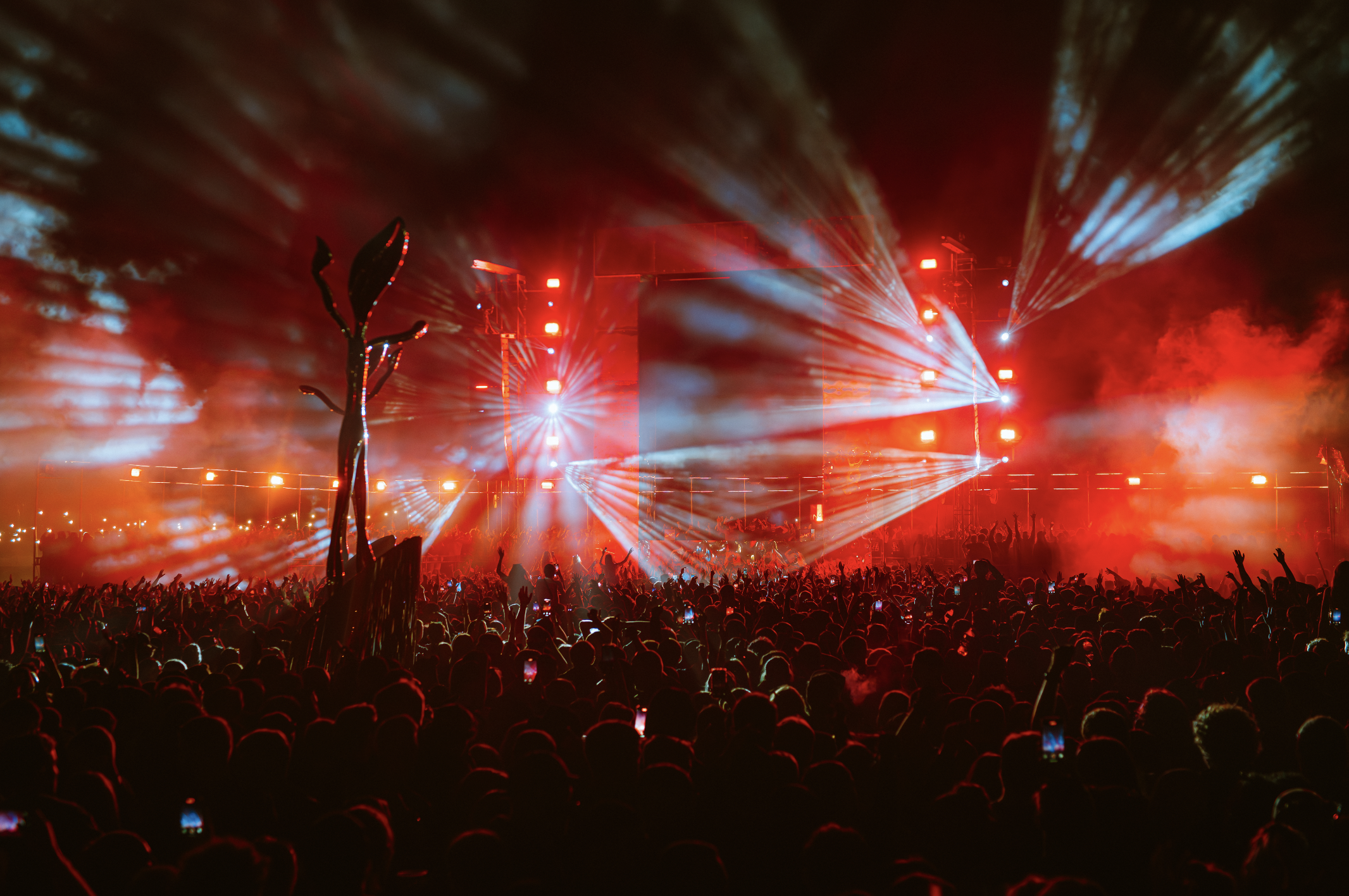 Crowd attending a concert or music festival at night with bright red and white stage lights, smoke effects, and silhouettes of people raising their hands.