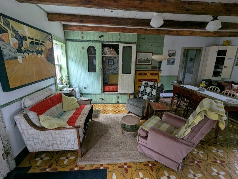 Rustic living room at Herdsman House with exposed beams, hand-painted Mennonite tile floors, retro furniture, and built-in green shelving.