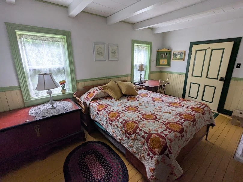 Cozy vintage-style bedroom with queen-sized bed, colorful quilt, and writing desk in the corner. Features mustard yellow painted wood floors, white walls with green trim, sheer-curtained windows, botanical wall art, and exposed ceiling beams. Include