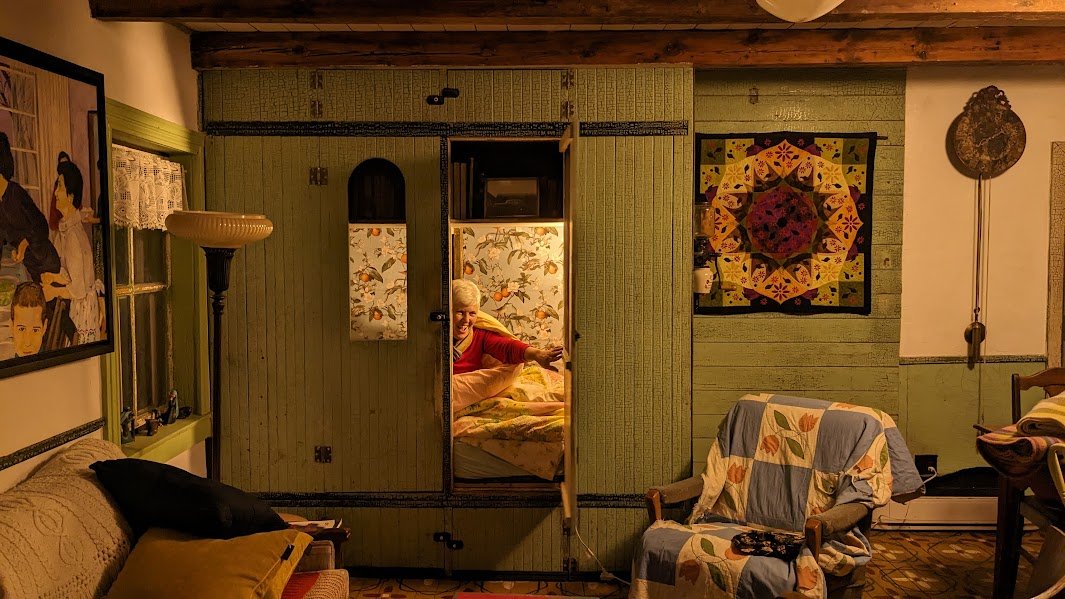 Bedstee-style sleeping nook in the living room at Herdsman House, built into a green-painted cabinet with paneled doors. Features orange bedding, yellow pillows, bird-and-foliage wallpaper, and built-in bookshelf. Restored Mennonite hand-painted floo