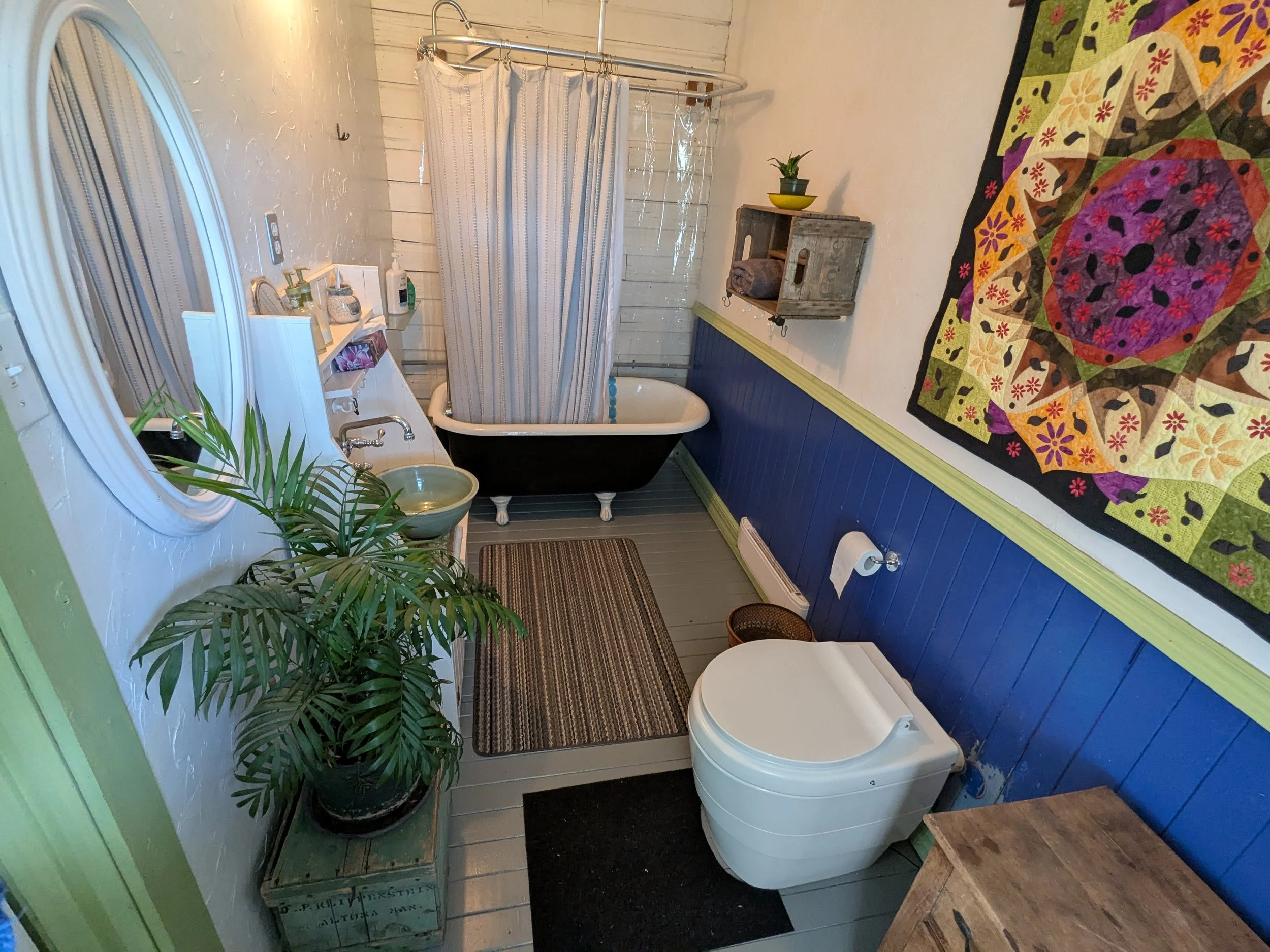 Eclectic bathroom with black clawfoot tub, green basin sink, and modern white composting toilet. Features blue and white walls with green trim, colorful quilted wall art, rustic shelf decor, potted plant on green stand, and patterned ceiling—blending