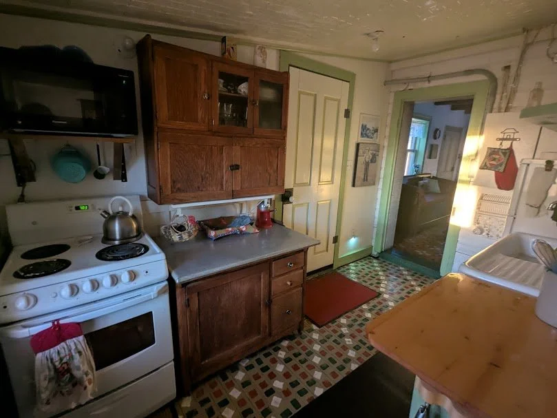 Cozy galley kitchen at Herdsman House with restored Mennonite tile floors, white gas stove, red coffee maker, and vintage wood cabinets.