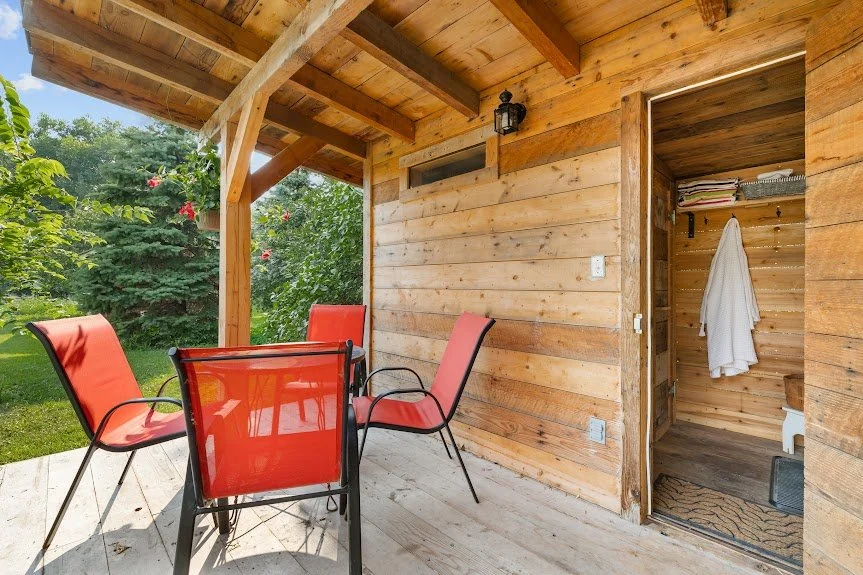 Covered porch of rustic sauna cabin with red cushioned chairs and glass-top table, surrounded by forest greenery at the Herdsman House retreat.