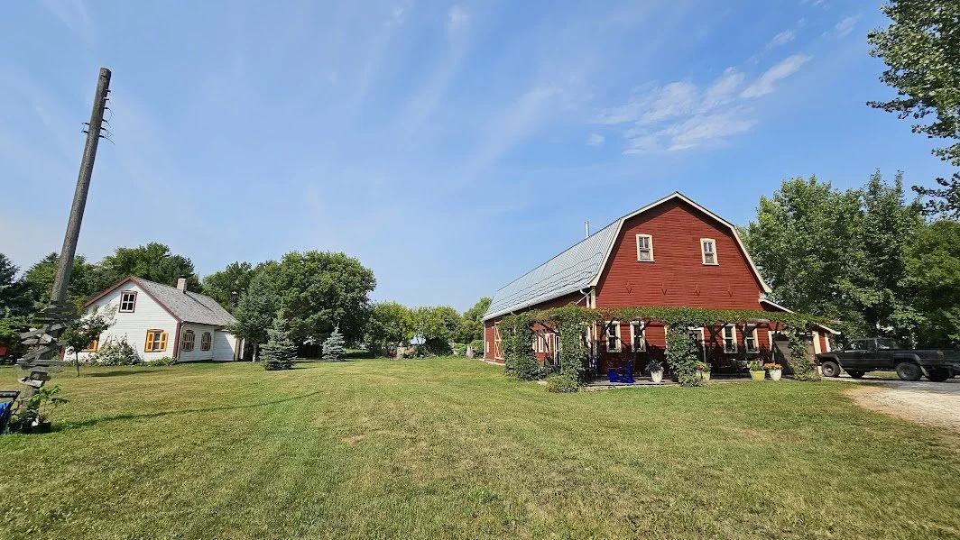 Red barn-style building with gray metal roof and covered porch, used for artist productions and concerts at the Herdsman House retreat in rural Manitoba.