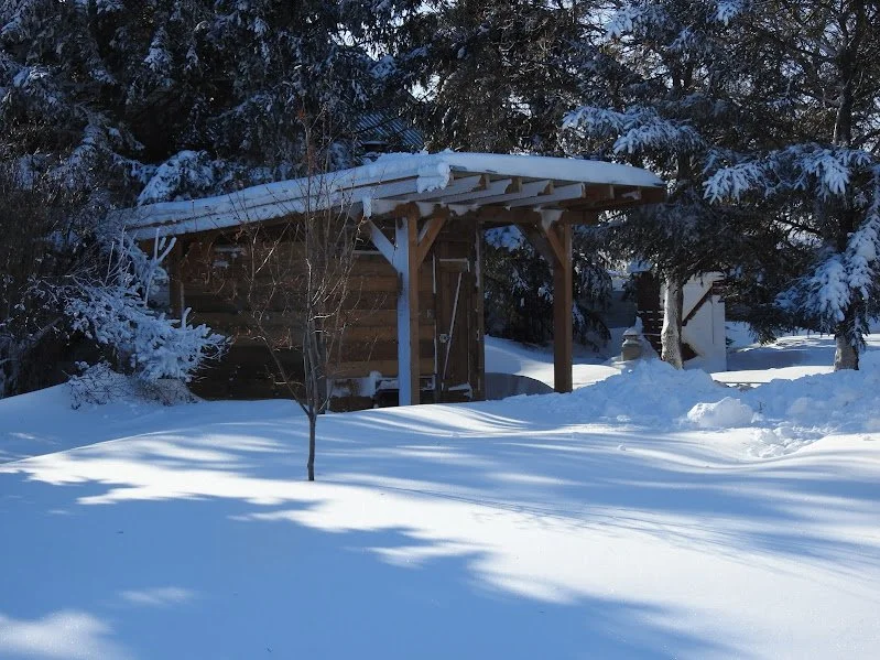 Nestled among snow-laden evergreens, this rustic wooden sauna cabin offers a serene escape for guests of the Herdsman House artist retreat. The slanted roof is blanketed in fresh snow, and the surrounding forest creates a peaceful, secluded atmospher