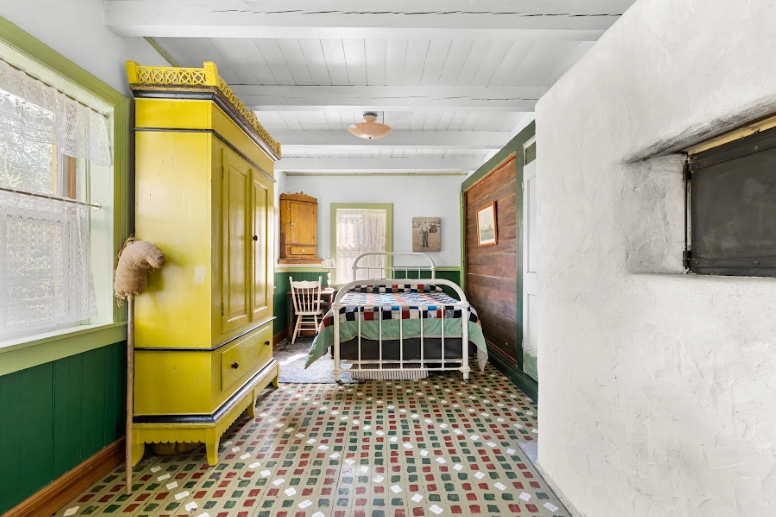 Warm, vintage-style bedroom with a white metal bed and patchwork quilt, set against wood-paneled walls. A cheerful yellow armoire stands beside restored hand painted Mennonite floor tiles in red, green, and white. Natural light spills through lace cu