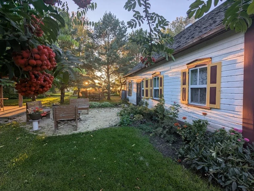 This idyllic outdoor scene features a picturesque white house with vibrant yellow and red trim, nestled among a flourishing garden of flowering plants and green shrubs. A cozy patio with wooden chairs and a table invites relaxation on the lush green 