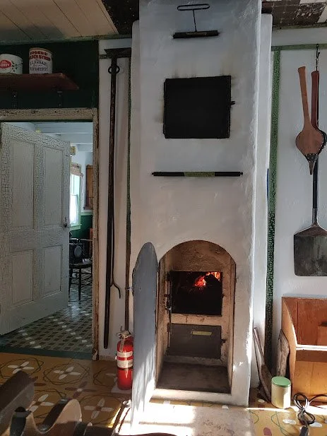 Traditional wood-burning stove built into a white plaster wall, with black metal doors used for cooking and heating the home. A shelf holds lit candles and a small figure, while red and white stockings hang above a cream-colored cabinet. Restored han