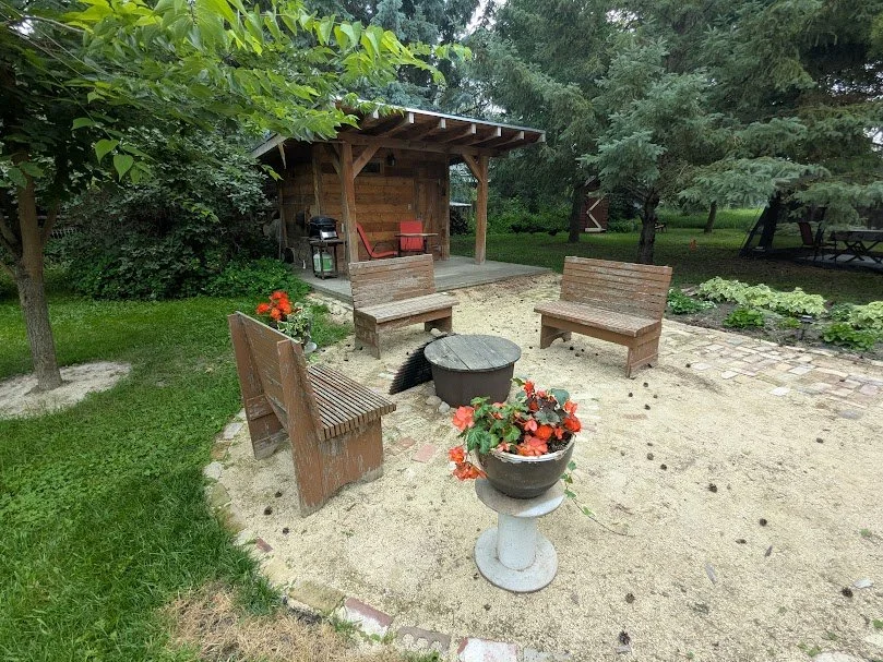 Cozy backyard fire pit with rustic wooden benches, red flower centerpiece, outdoor grill pavilion, and vegetable garden surrounded by trees and lawn.