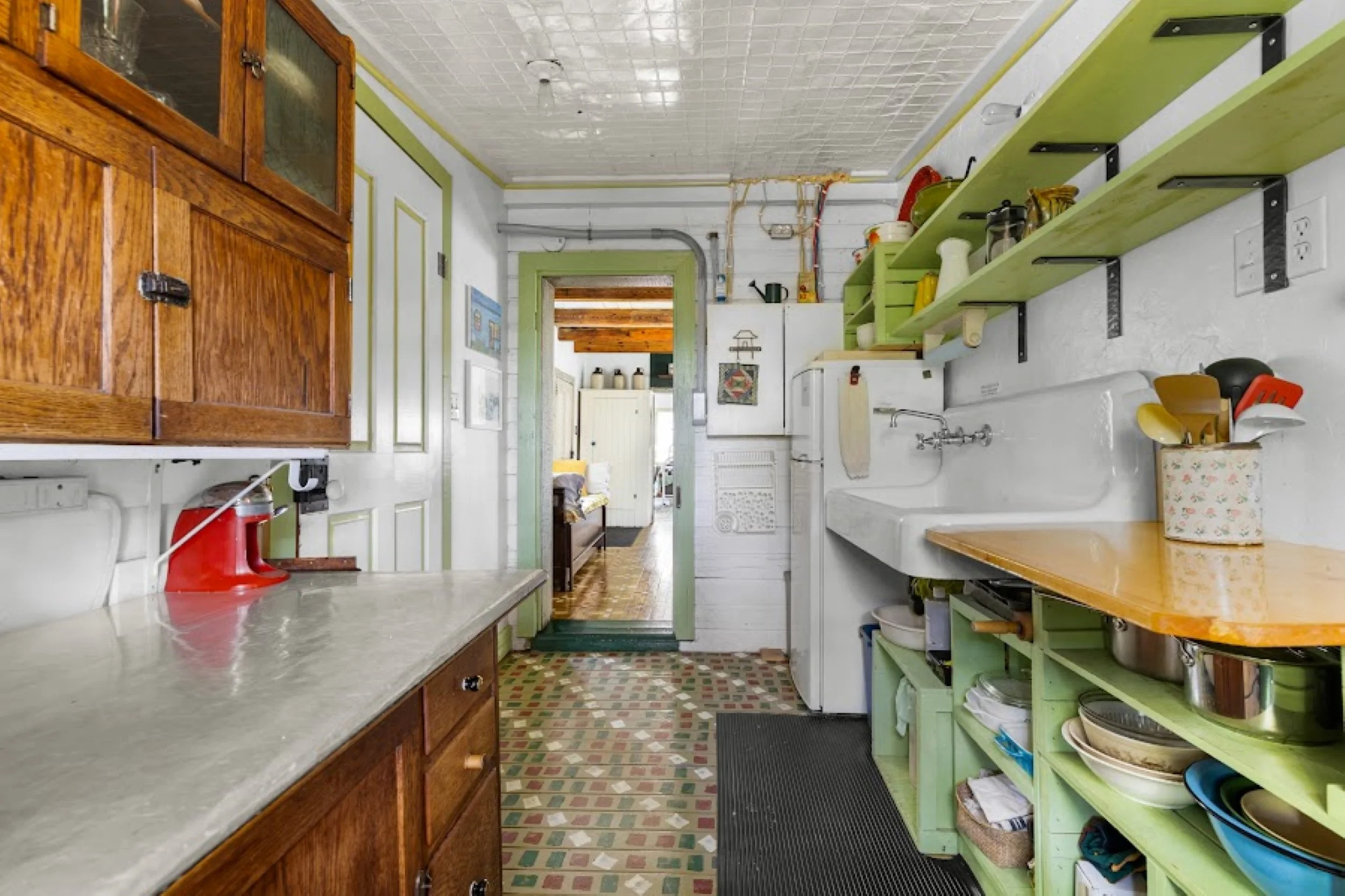 Vintage kitchen with a white porcelain sink, green open shelving, and restored hand painted Mennonite floor tiles in a vibrant mosaic of red, green, and white. Natural light filters through a central window, illuminating pressed tin ceilings, a cheer