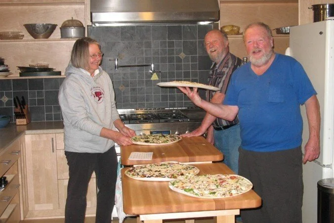 Collaboration, Three people in a kitchen, two men and one woman, sharing and presenting pizzas on a wooden table.