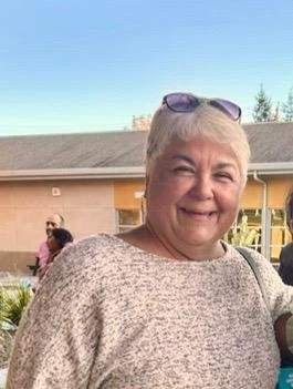 A smiling older woman with short gray hair, wearing sunglasses on her head and a light-colored sweater, standing outdoors with a yellow building and clear blue sky in the background.