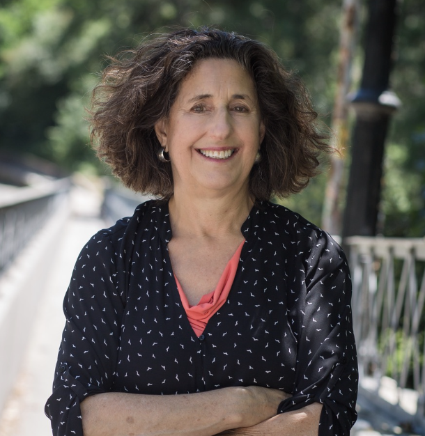 A smiling middle-aged woman with curly brown hair, wearing a black blouse with white patterns and an orange top underneath, standing outdoors on a sunny day with trees and a walkway in the background. Kathryn McCamant cohousing development consultant