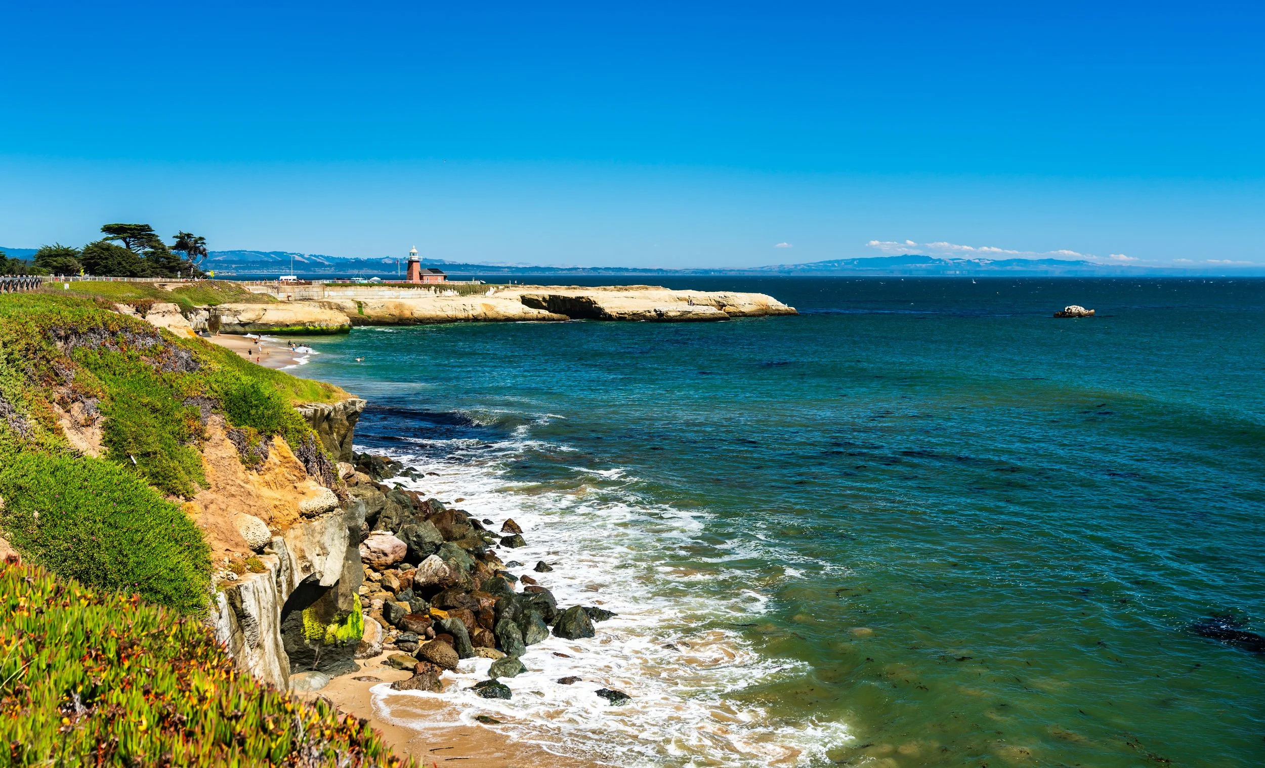 Coastal scene in Santa Cruz, California, with rocky cliffside and sandy beach, lighthouse in the distance under a clear blue sky, calm ocean water.
