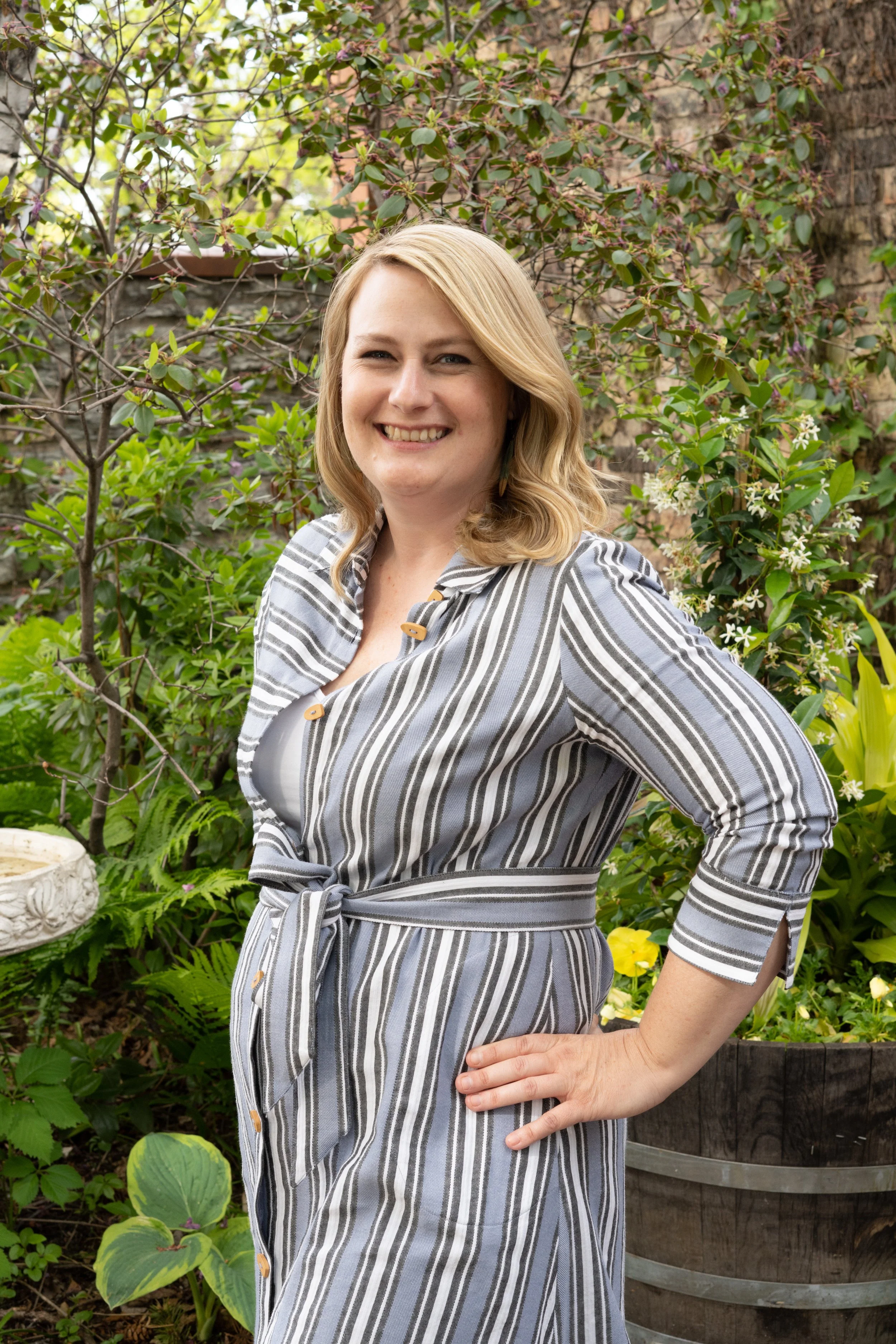 A woman with blonde hair smiling and standing outdoors in a garden with green foliage and blooming flowers. Christina Marshall with Coho Collab.  Cohousing membership and marketing