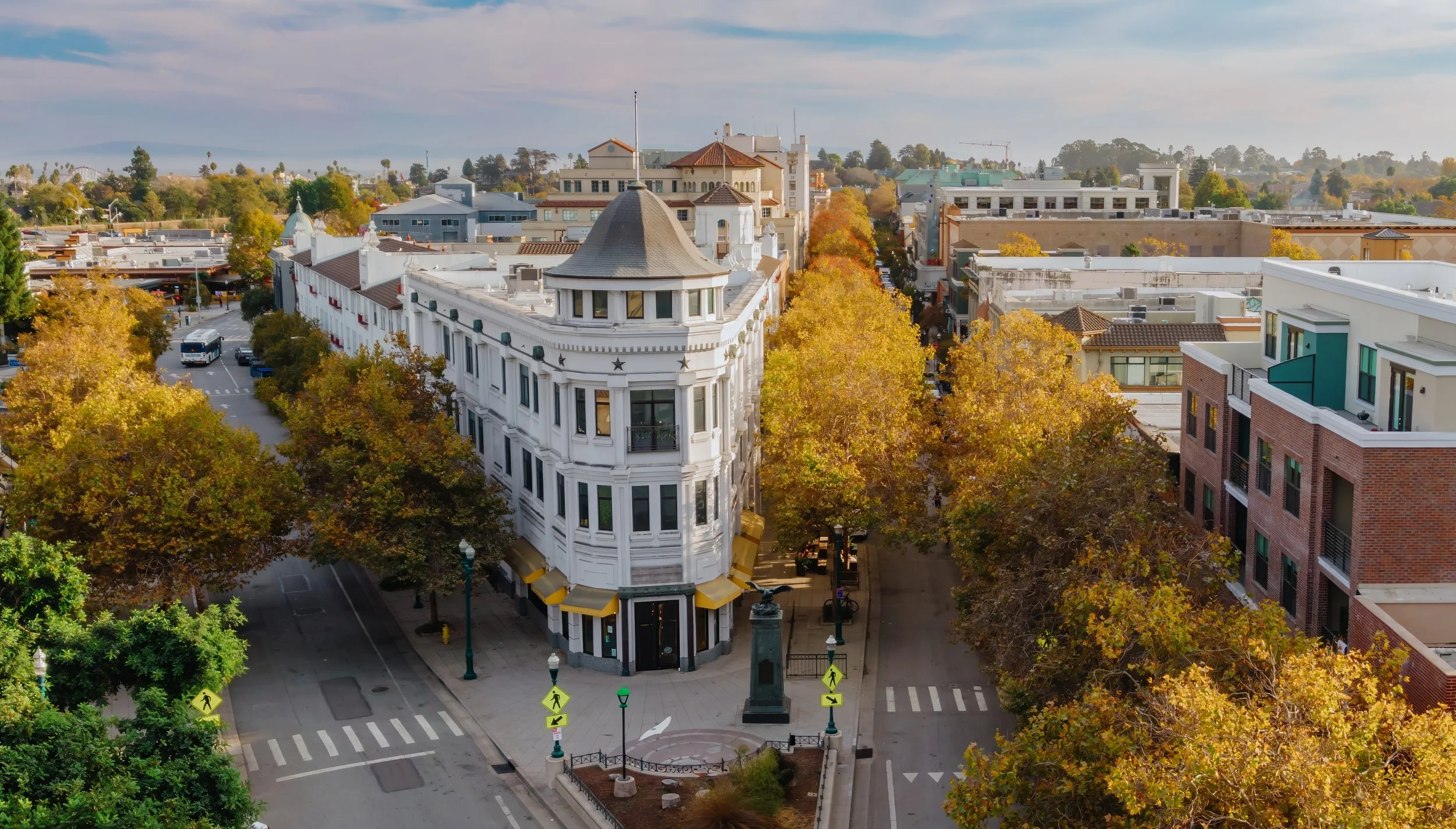 Downtown Santa Cruz.  A city street intersection with trees displaying autumn foliage, a white historic building with a rounded tower corner, and surrounding modern buildings, under a cloudy sky.