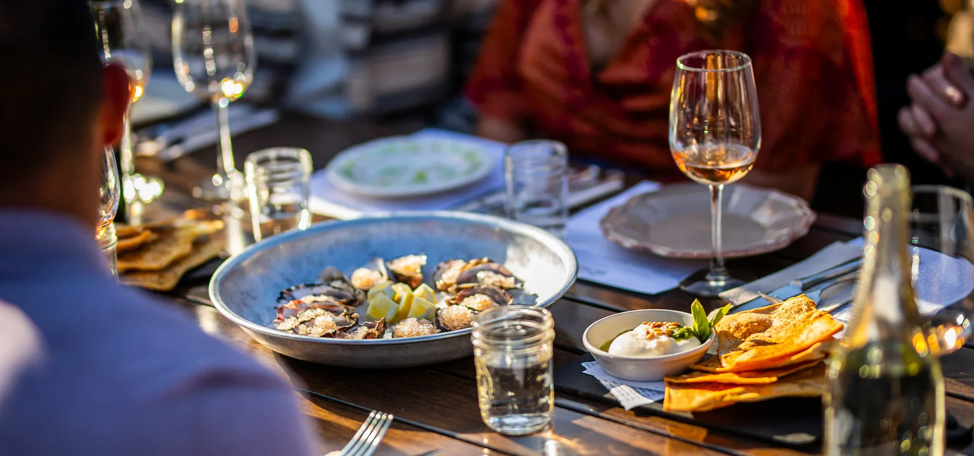 A dinner table set with dishes including a plate of oysters, a bowl of dip or sauce, and a serving of sliced bread or crackers. There are glasses of wine, water, and other drinkware, with a background of people dining outdoors.